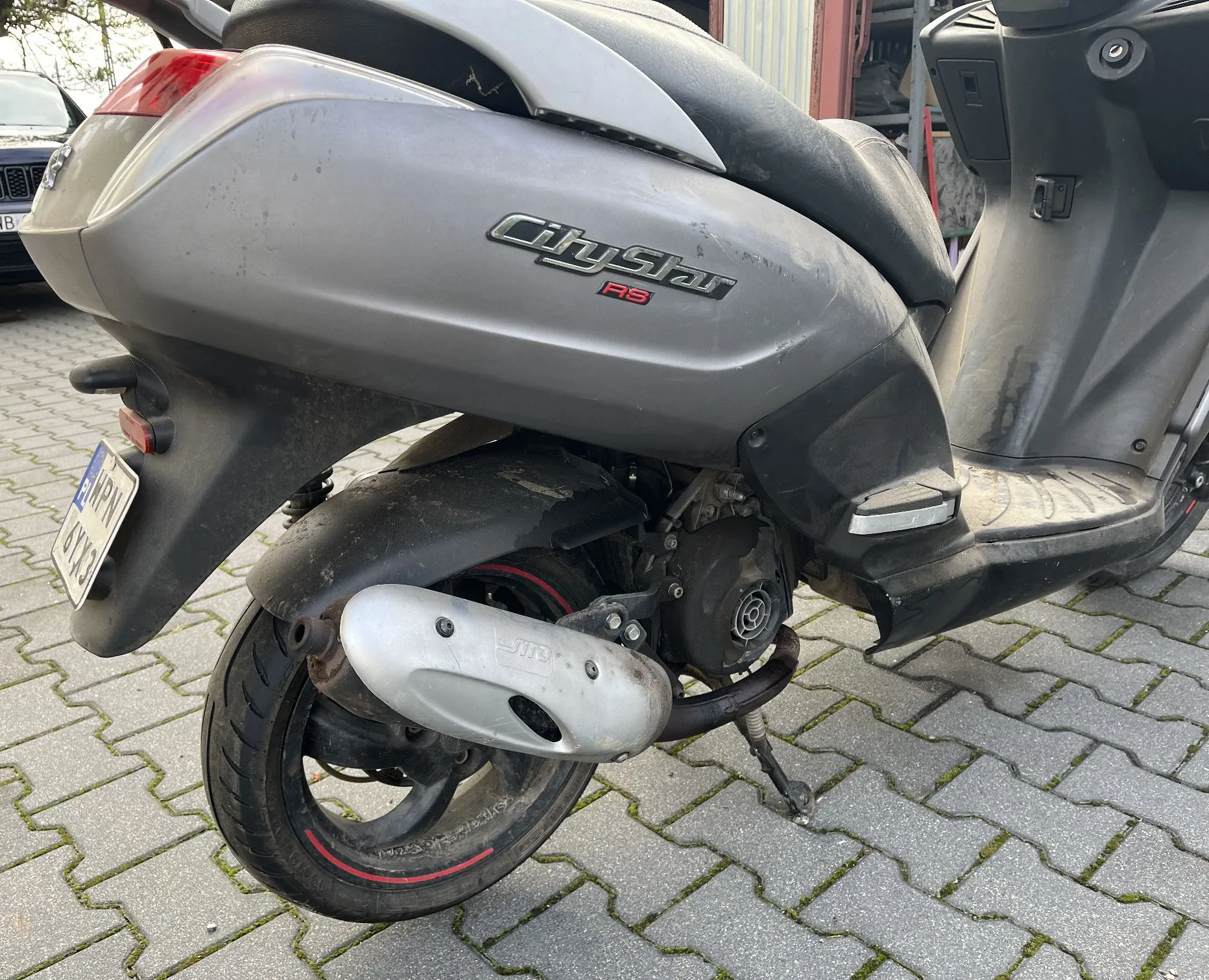 Tylna część szarego skutera zaparkowanego na kostce brukowej. Na boku skutera znajduje się napis „CityStar RS”. Widoczne jest tylne koło z czarną oponą z czerwoną linią na feldze, srebrny tłumik wydechu oraz fragment tablicy rejestracyjnej.
