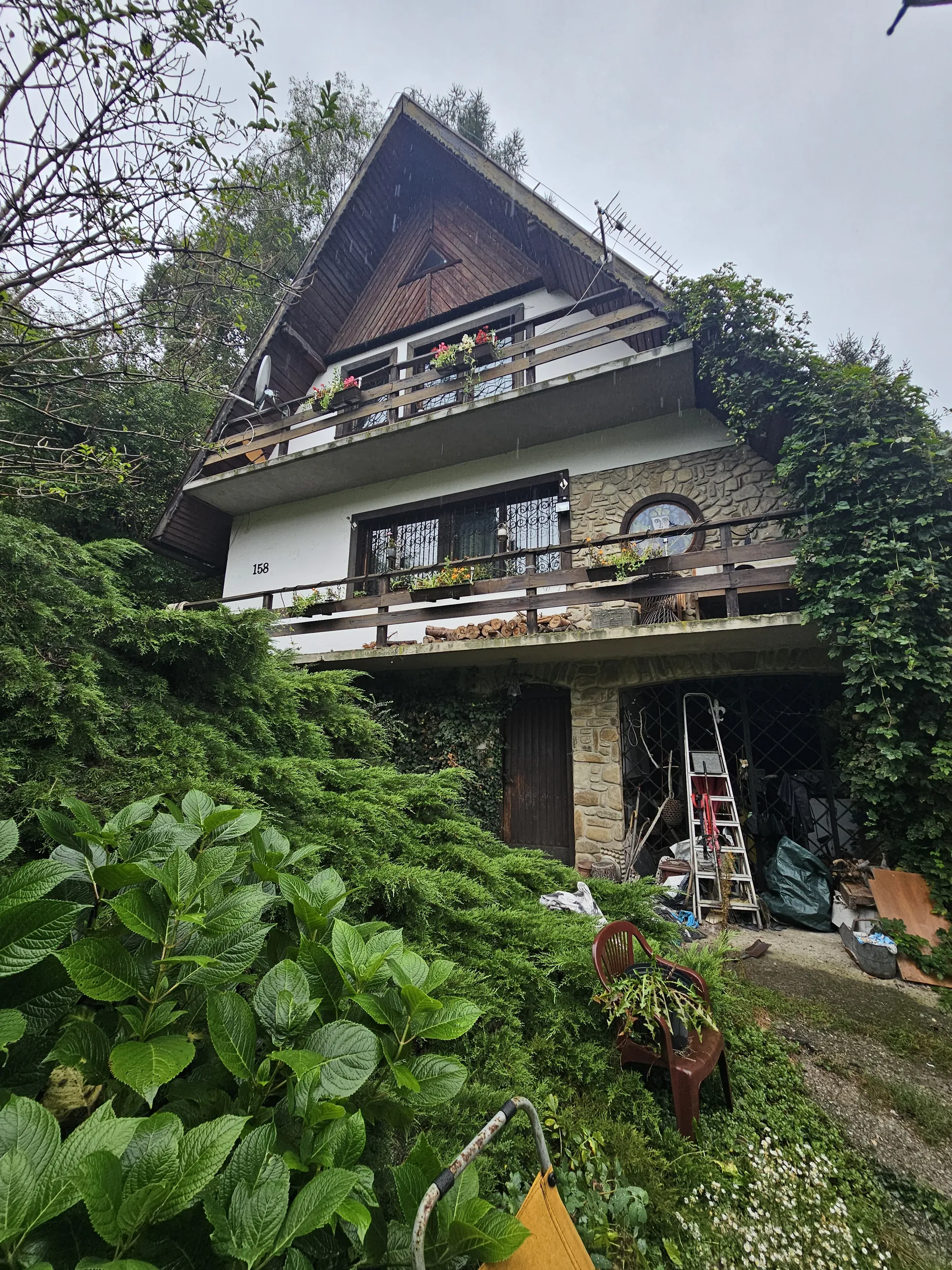 Dom z kamiennej i drewnianej elewacji, numer 158 na białej ścianie. Balkon z kwiatami, zarośla i krzewy wokół. Widoczne krzesła, drabina oraz różne przedmioty przy wejściu. Niebo zachmurzone.