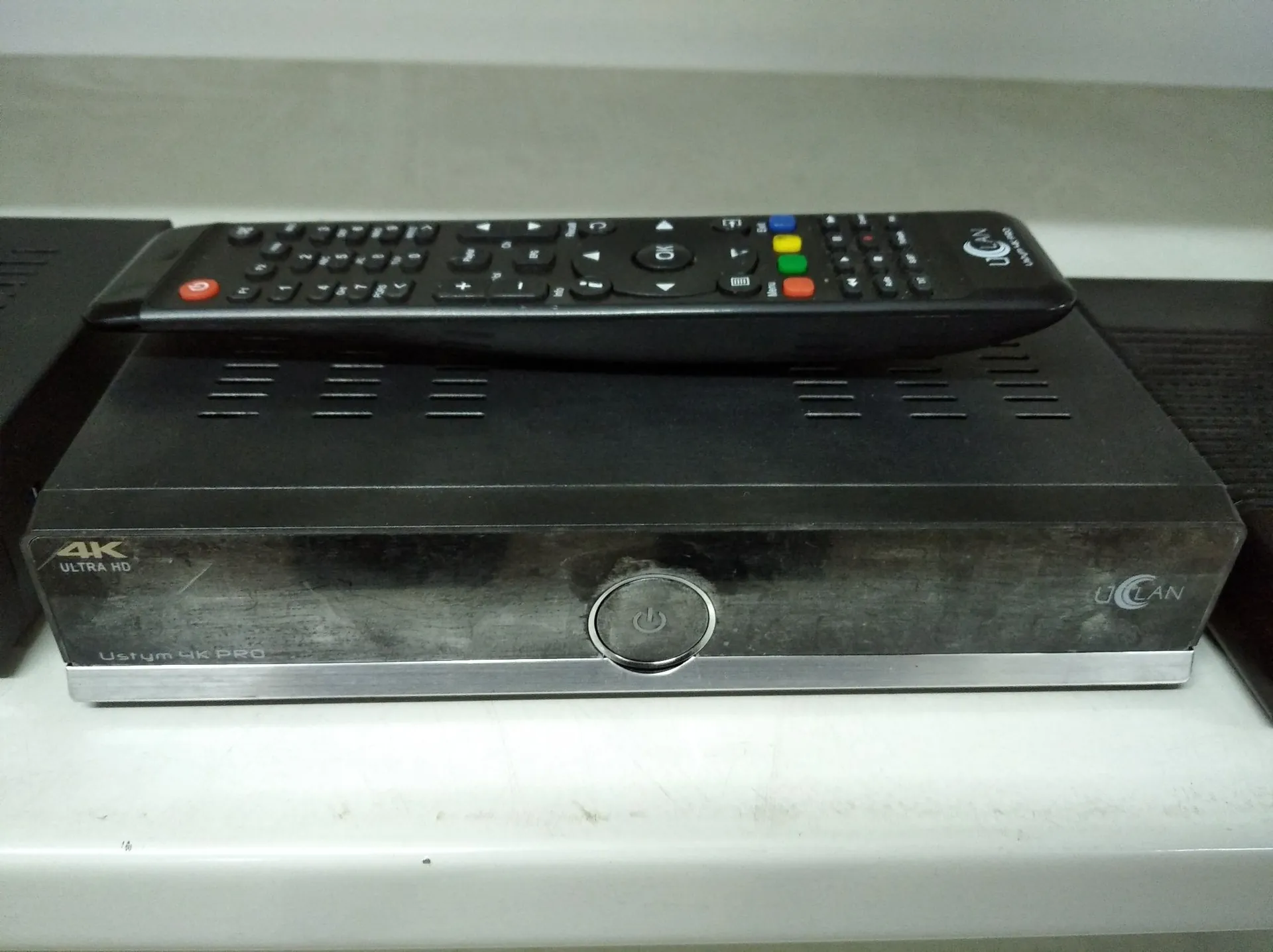 Czarny dekoder telewizyjny z napisem '4K ULTRA HD' po lewej stronie, 'Ucan' po prawej oraz 'Ustym 4K PRO' na dolnej srebrnej listwie. Na urządzeniu znajduje się okrągły przycisk zasilania. Na górze dekodera leży czarny pilot z wieloma przyciskami, w tym kolorowymi przyciskami sterowania oraz logo 'Ucan'.