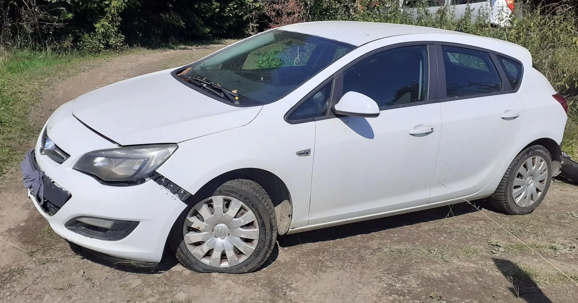 Biały samochód osobowy typu hatchback zaparkowany na piaszczystym podłożu. Pojazd jest uszkodzony w przedniej części: brakuje fragmentu przedniego zderzaka po prawej stronie (patrząc od przodu), a maska jest lekko uchylona. Samochód stoi bokiem do obiektywu, w tle widoczna jest zieleń krzewów i drzew.
