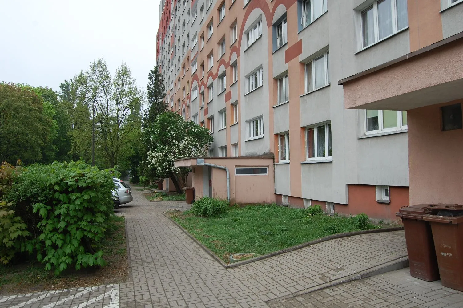 Widok na wielopiętrowy blok mieszkalny z jasnoszarą i pomarańczową elewacją. Przed budynkiem znajduje się chodnik z kostki brukowej, pas zieleni z trawą oraz gęste zielone krzewy po lewej stronie. W oddali widać zaparkowany srebrny samochód i drzewa. Po prawej stronie, przy wejściu do budynku, stoją dwa brązowe pojemniki na odpady.