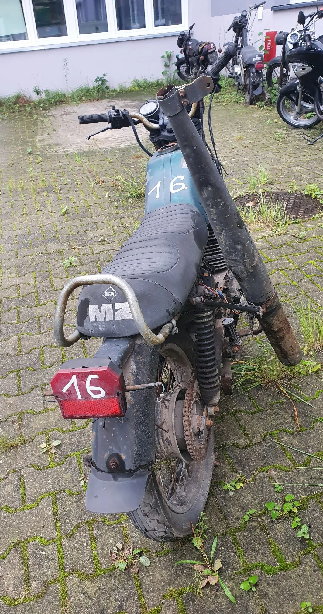 Widok z tyłu na stary, niebieski motocykl marki MZ, zaparkowany na kostce brukowej porośniętej mchem. Na baku oraz na tylnej lampie widnieje biała liczba 16. Motocykl posiada czarne siedzenie, metalowy stelaż z napisem MZ oraz wysoką, zardzewiałą rurę wydechową biegnącą wzdłuż prawej strony. W tle widoczne są inne motocykle oraz biała ściana budynku z oknami.