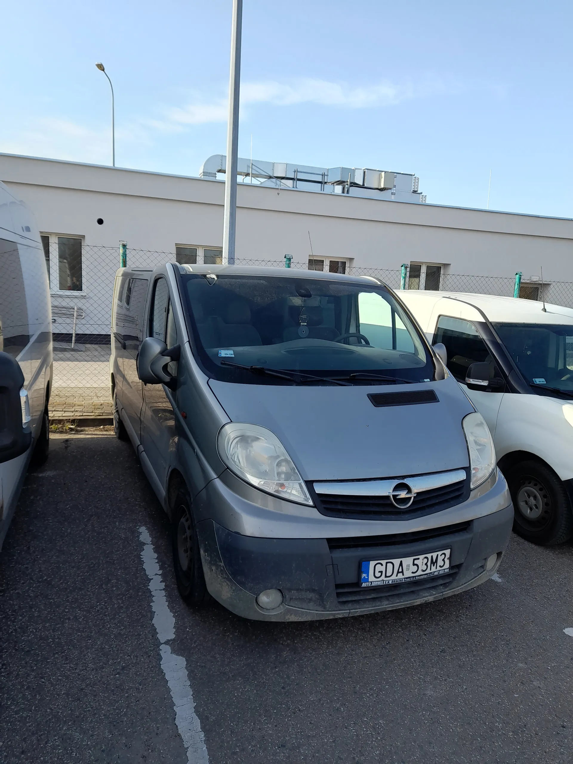 Trzy samochody zaparkowane na asfaltowym placu parkingowym. Na pierwszym planie szary samochód osobowy z tablicą rejestracyjną GDA 53M3, za nim biały samochód. W tle budynek przemysłowy lub biurowy z oknami i siatką ochronną. Niebo jest jasne z kilkoma chmurami. Widoczne są również latarnie uliczne. Tekst na tablicy rejestracyjnej: GDA 53M3 oraz mały napis PL na niebieskim pasie.