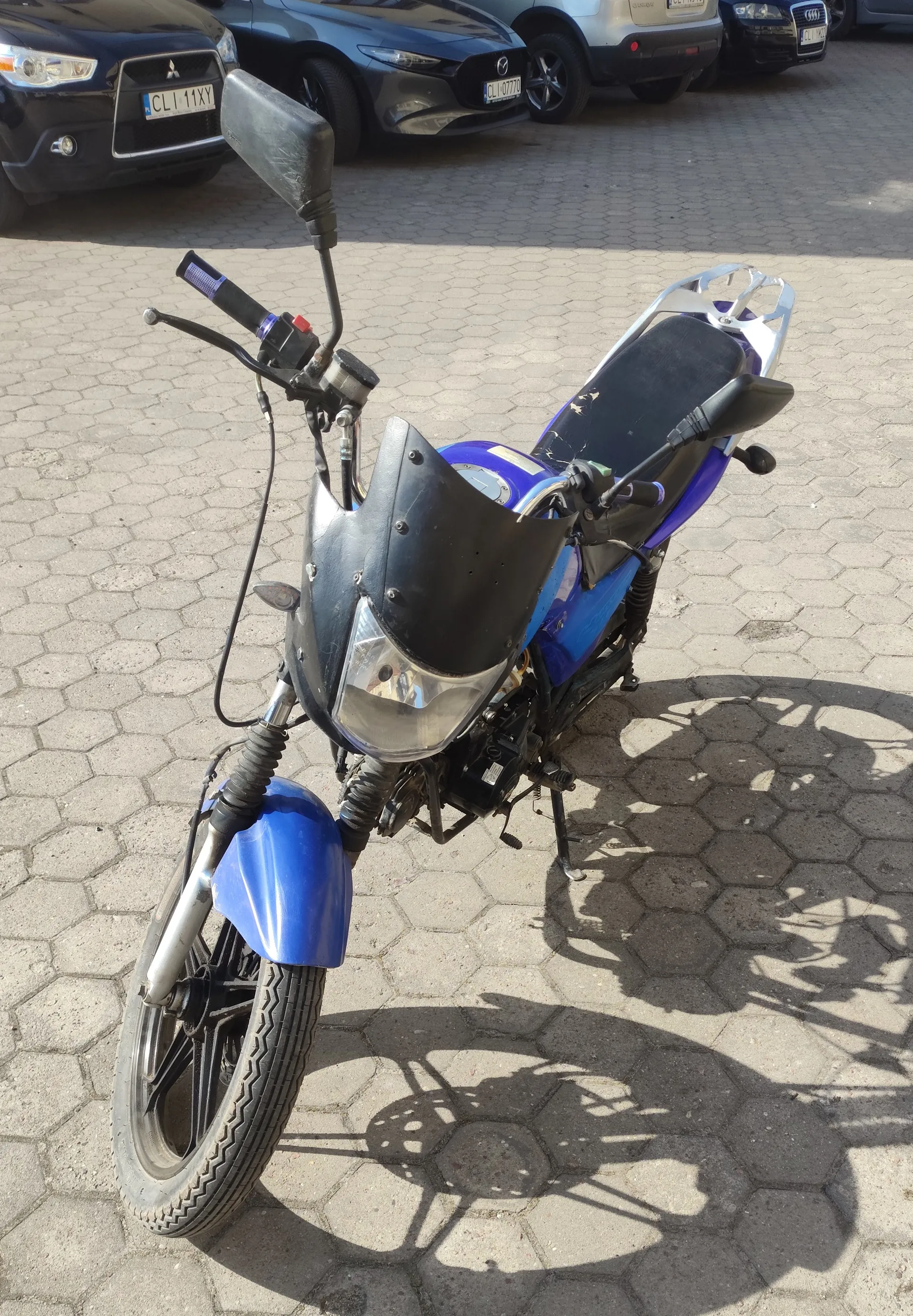 Niebiesko-czarny motocykl z czarnym siedzeniem, kierownicą z lusterkami i reflektorem, zaparkowany na brukowanej powierzchni z sześciokątnymi płytami. W tle widoczne są samochody: czarny Mitsubishi z tablicą rejestracyjną CLI 11XY, szary Mazda z tablicą CLI 0770, srebrny SUV oraz czarny Audi. Cień motocykla na ziemi.