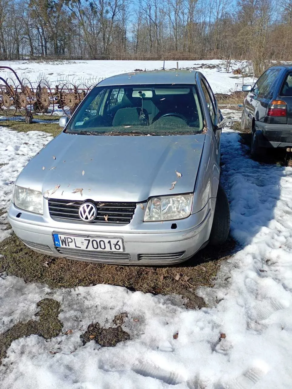 Srebrny samochód marki Volkswagen z tablicą rejestracyjną WPL 70016, zaparkowany na obszarze z resztkami śniegu i trawą. W tle widoczne są suche drzewa, rolnicze narzędzia oraz częściowo widoczny czarny samochód po prawej stronie. Na przedniej szybie i masce znajdują się suche liście.