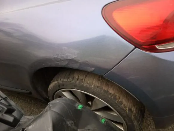 Zbliżenie na tylną część szarego samochodu z widocznym uszkodzeniem karoserii w okolicy nadkola. Czerwone światło tylnego reflektora, brudna opona z aluminiową felgą oraz czarny plastikowy element z zielonymi akcentami w pierwszym planie. Brak czytelnego tekstu.