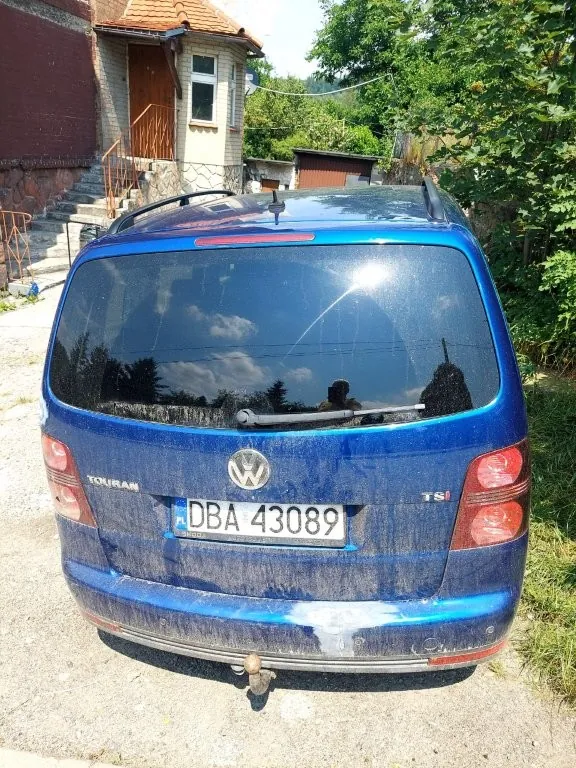 Tylny widok niebieskiego samochodu marki Volkswagen Touran z silnikiem TSI. Pojazd jest zabrudzony pyłem i błotem, szczególnie w dolnej części nadwozia. Na klapie bagażnika znajduje się logo Volkswagena oraz tablica rejestracyjna o numerze DBA 43089. Samochód posiada hak holowniczy. W tle widoczny jest fragment budynku z cegły i dachówką oraz zielone drzewa.