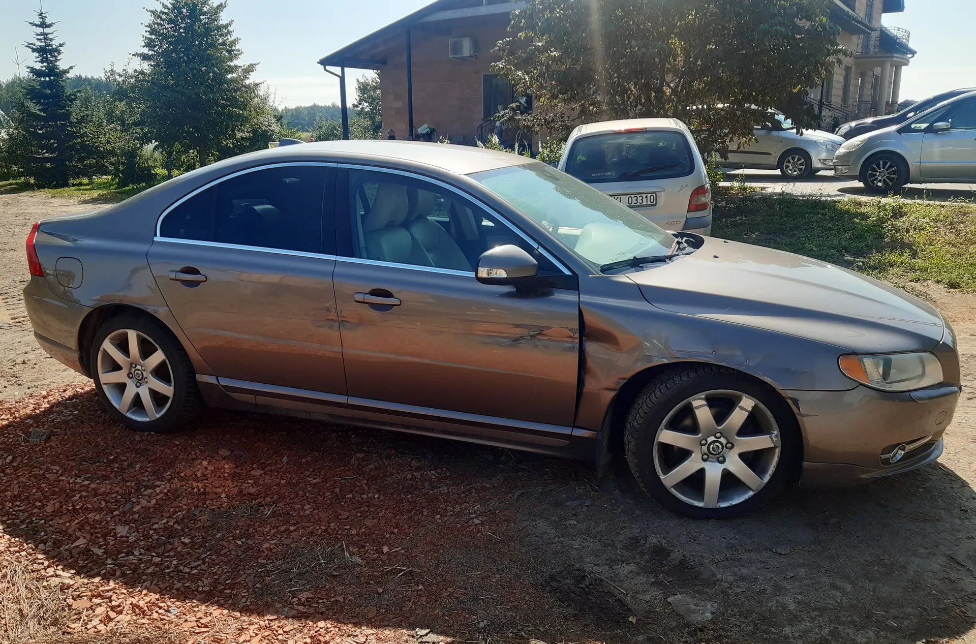 Brązowy samochód osobowy typu sedan zaparkowany na piaszczystym podłożu. Pojazd ma widoczne uszkodzenia karoserii w postaci wgnieceń na przednich i tylnych drzwiach od strony pasażera. W tle widoczne są inne zaparkowane samochody, dom oraz drzewa pod jasnym, słonecznym niebem.