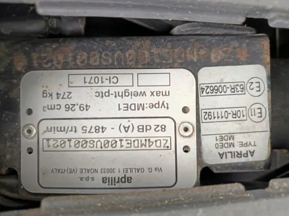 Dwa metalowe tablice identyfikacyjne z widocznym tekstem. Górną tablicę zajmuje napis 'aprilia s.p.a.', poniżej adres 'Via G. GALILEI 1 30033 NOALE (VE) ITALY', numer seryjny 'ZD4MDE100US001021', dane techniczne: '82 dB (A) - 4875 tr/min', 'type:MDE1', '49,26 cm3', 'max weight-ptc 274 kg' oraz kod 'CI-1071'. Dolna tablica zawiera napis 'APRILIA', 'TYPE MDE0 MDE1', oznaczenia 'E11 10R-011192' i 'E2 63R-006624'. Tablice zamocowane są na ciemnym, metalowym podłożu z widocznymi śrubami.