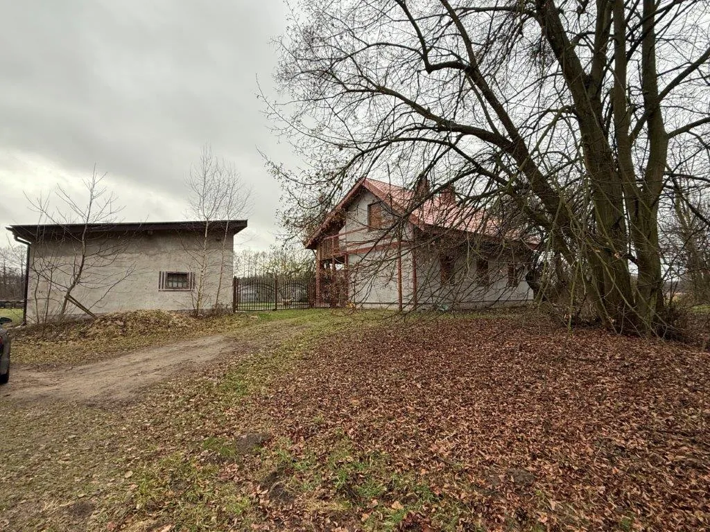 Zdjęcie przedstawiające dwa budynki na posesji w pochmurny dzień. Po lewej stronie znajduje się niski, prostokątny budynek gospodarczy z szarymi ścianami i płaskim dachem. Po prawej stronie stoi większy dom z czerwonym, spadzistym dachem i jasnymi ścianami. Pomiędzy budynkami widoczny jest metalowy płot. Przed budynkami znajduje się teren pokryty brązowymi, opadłymi liśćmi oraz fragmenty trawnika i piaszczysta droga. Po prawej stronie zdjęcia dominują duże, bezlistne drzewa o gęstych gałęziach.