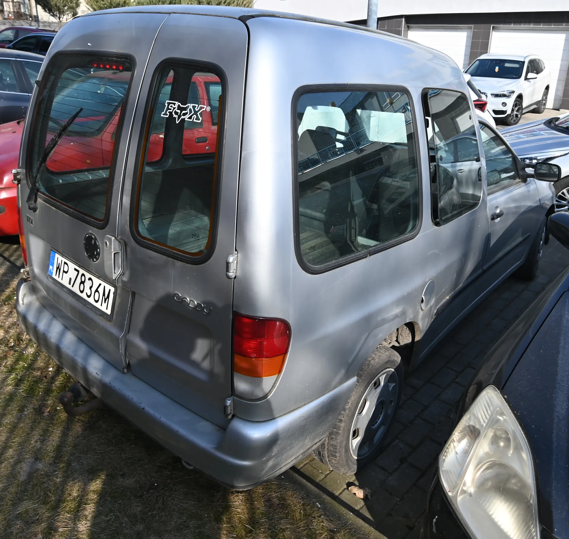 Srebrny samochód dostawczy Volkswagen Caddy zaparkowany na podwórku. Widoczny tył pojazdu z tablicą rejestracyjną WP.7836M oraz naklejką "FOX" na tylnej szybie. W tle inne samochody: czerwony, biały SUV i srebrny sedan. Podłoga z cegły i trawnik po lewej stronie. Budynki z białymi bramami garażowymi w tle.