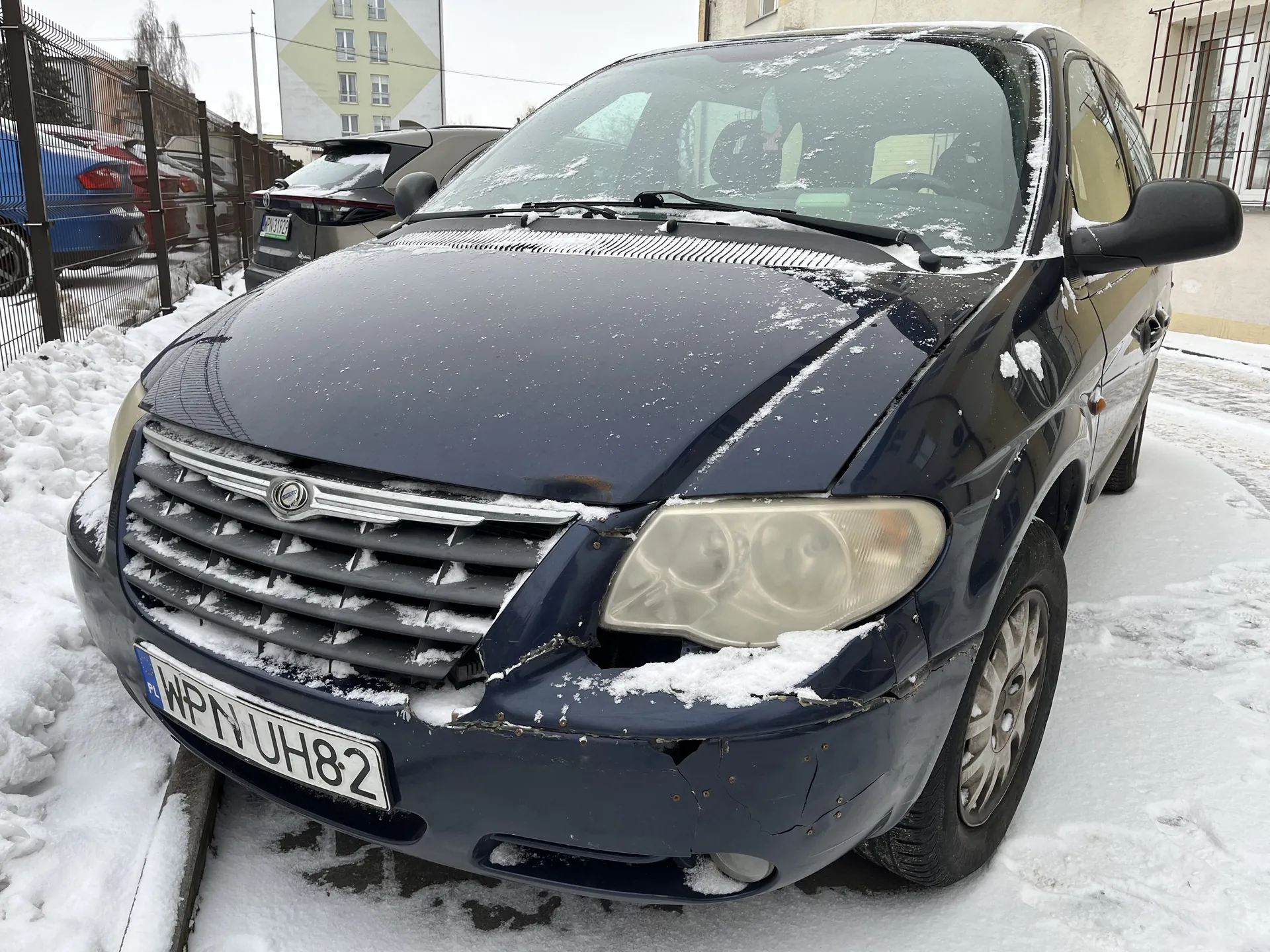 Ciemnoniebieski samochód z widocznymi uszkodzeniami przedniej części, w tym pękniętym zderzakiem i maską, pokryty warstwą śniegu. Tablica rejestracyjna WPN UH82. W tle stoją inne pojazdy, metalowe ogrodzenie oraz budynki mieszkalne. Śnieg pokrywa ziemię i powierzchnie samochodów, tworząc zimowy krajobraz.