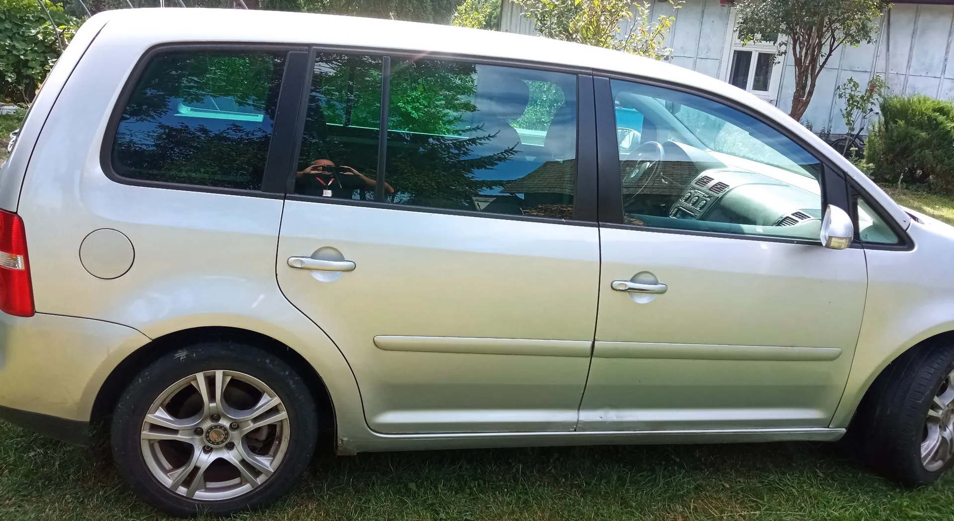 Srebrny samochód typu minivan zaparkowany na trawie, pokazany z profilu od strony pasażera. Widoczne są dwa drzwi, koła z aluminiowymi felgami oraz szyby, w których odbija się zieleń drzew.