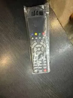 Zdjęcie przedstawia nowy pilot w przezroczystym opakowaniu zabezpieczającym, leżący na ciemnej powierzchni. Pilot posiada kolorowe przyciski (czerwony, zielony, żółty, niebieski), okrągły pad kierunkowy oraz inne przyciski funkcyjne. Na opakowaniu widoczny jest napis '150.00' umieszczony pionowo po prawej stronie obrazu.