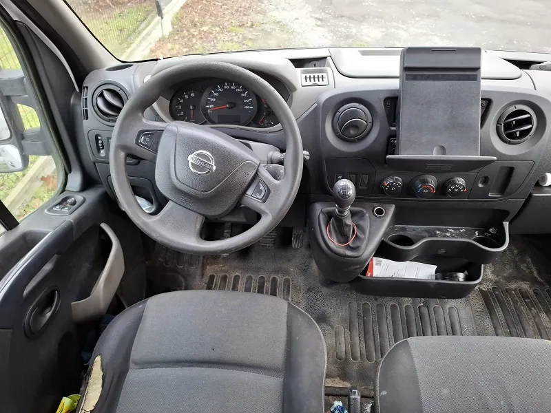 Wnętrze pojazdu z widocznym kierownicą marki Nissan, tablicą rozdzielczą z licznikami, dźwignią zmiany biegów, konsolą środkową z przyciskami i uchwytem na dokumenty. Widoczne są również przednie siedzenia, drzwi z otwartym oknem oraz zewnętrzny widok na trawnik i drogę. Podłoga pokryta jest brudnymi dywanikami.