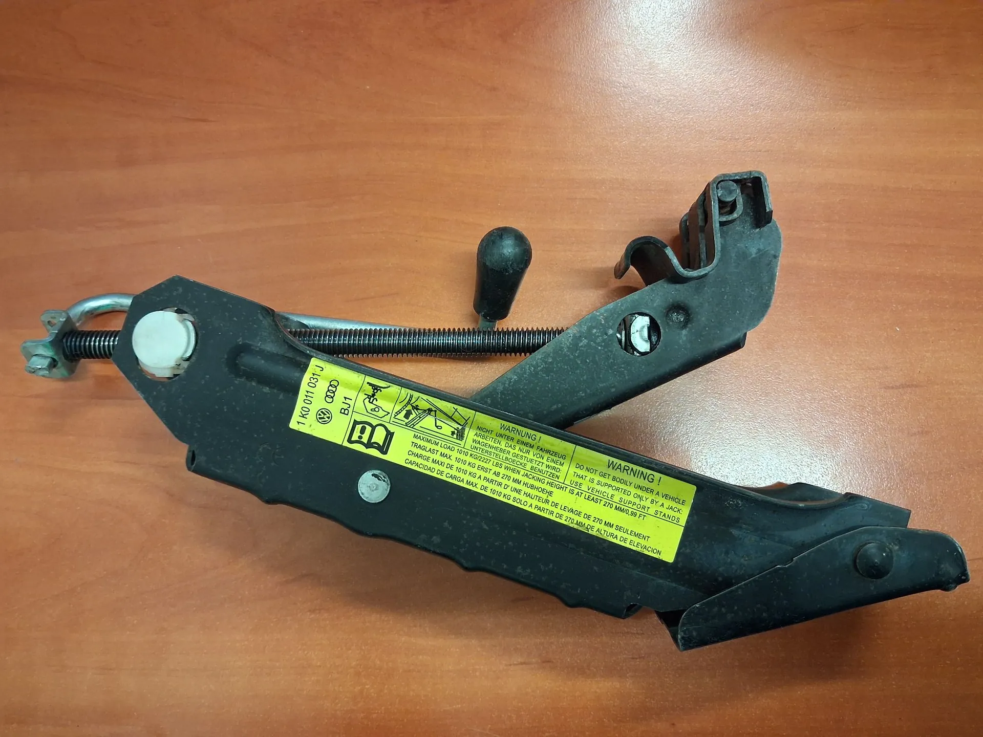 Metalowy mechanizm podnośnika samochodowego w kolorze czarnym, leżący na brązowej, drewnianej powierzchni. Urządzenie posiada rączkę z czarnym uchwytem oraz żółtą naklejkę ostrzegawczą z tekstem w kilku językach, w tym angielskim: 'WARNING! DO NOT GET BODY UNDER A VEHICLE'. Na obudowie widnieje również oznaczenie '1K0 011 031J' oraz logo marki Volkswagen.