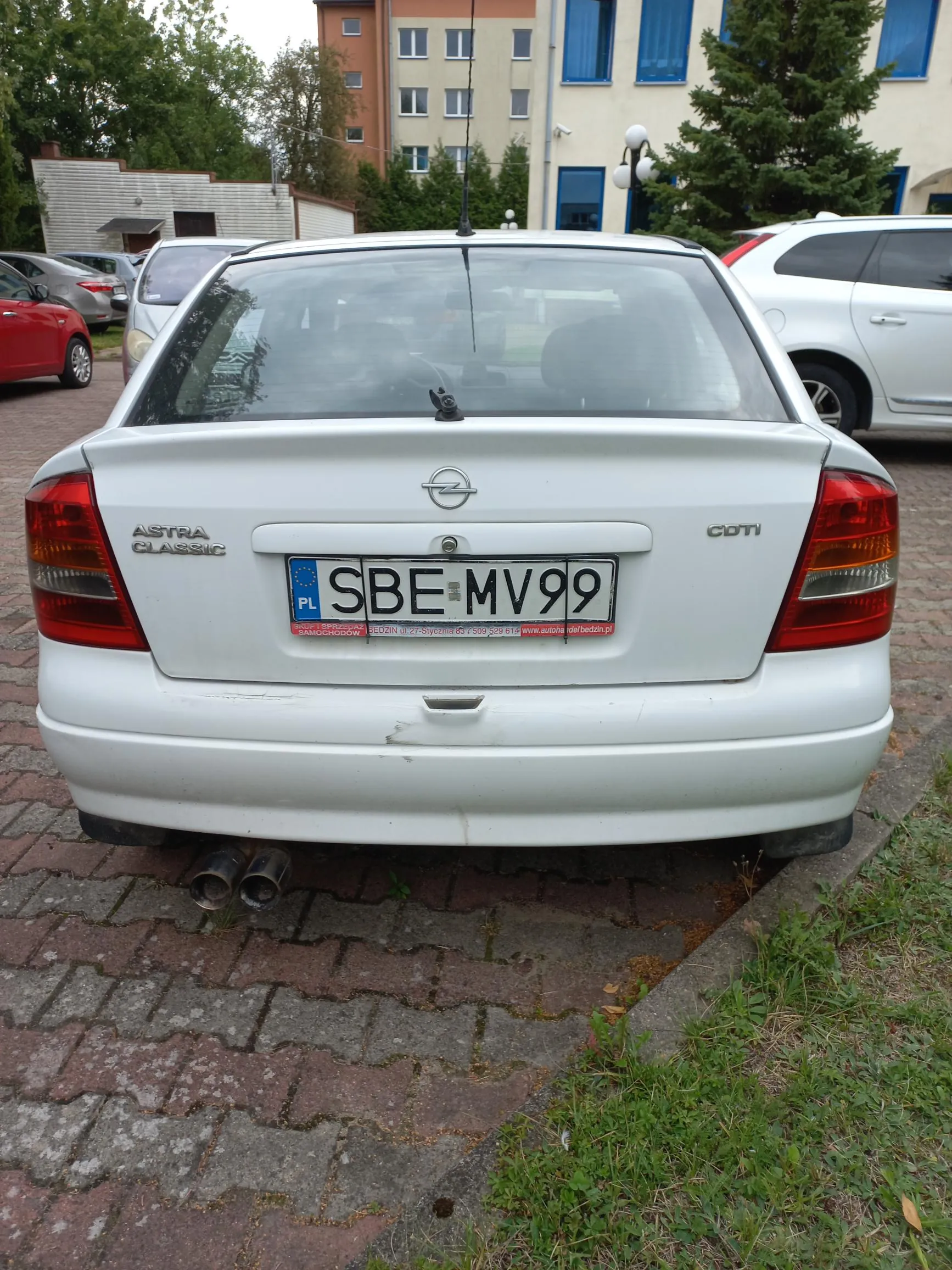 Tylny widok białego samochodu marki Opel Astra Classic CDTI zaparkowanego na kostce brukowej. Samochód ma polską tablicę rejestracyjną o numerze SBE MV99. Po lewej stronie widoczny jest fragment czerwonego auta, a po prawej biały samochód typu kombi. W tle znajduje się wielopiętrowy budynek w kolorze żółtym i beżowym oraz zielone drzewa.