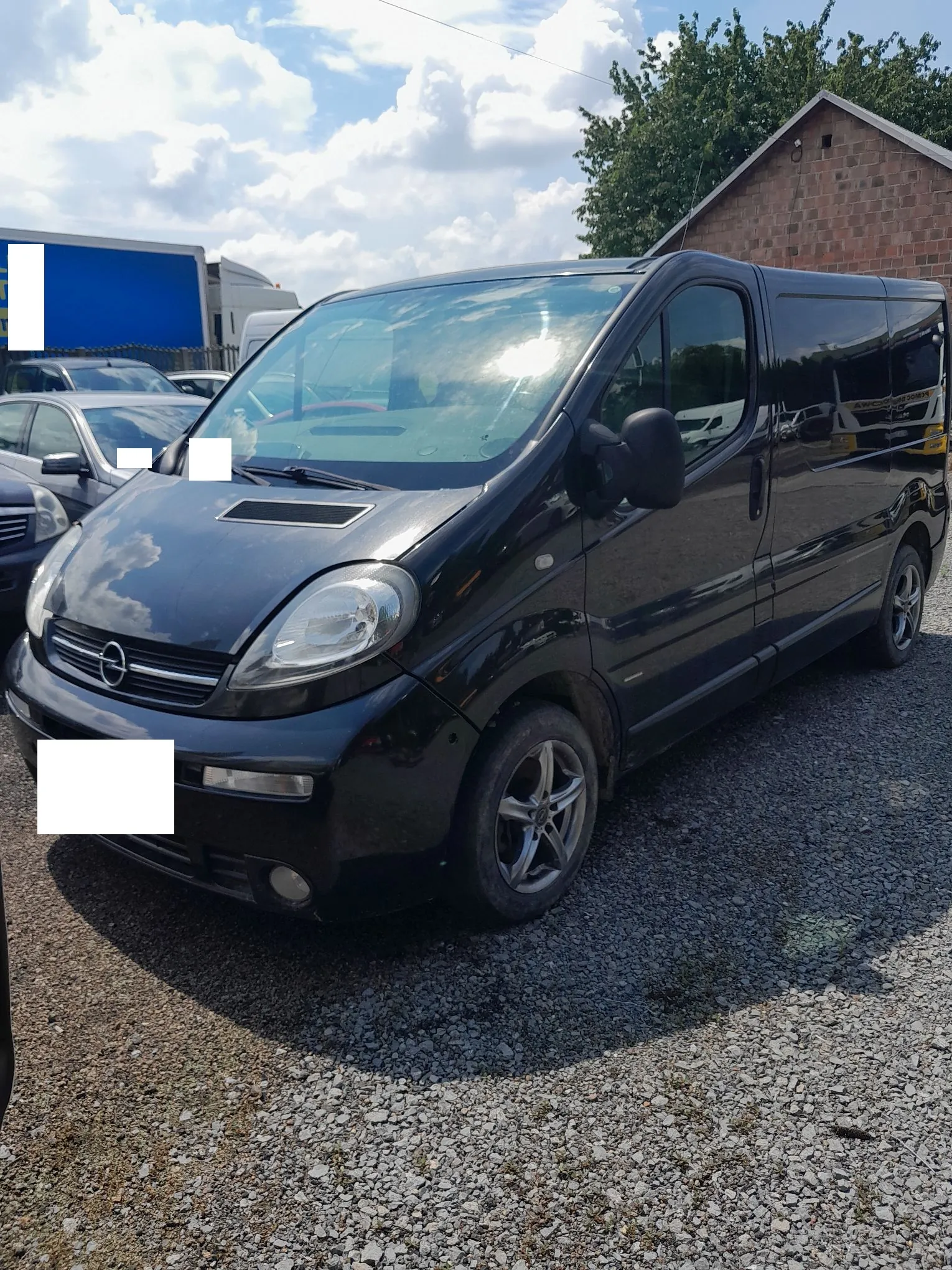 Czarny samochód dostawczy marki Opel zaparkowany na żwirowym placu. Pojazd jest widoczny z przodu i z boku. W tle znajduje się niebieska ciężarówka oraz ceglany budynek pod częściowo zachmurzonym niebem.