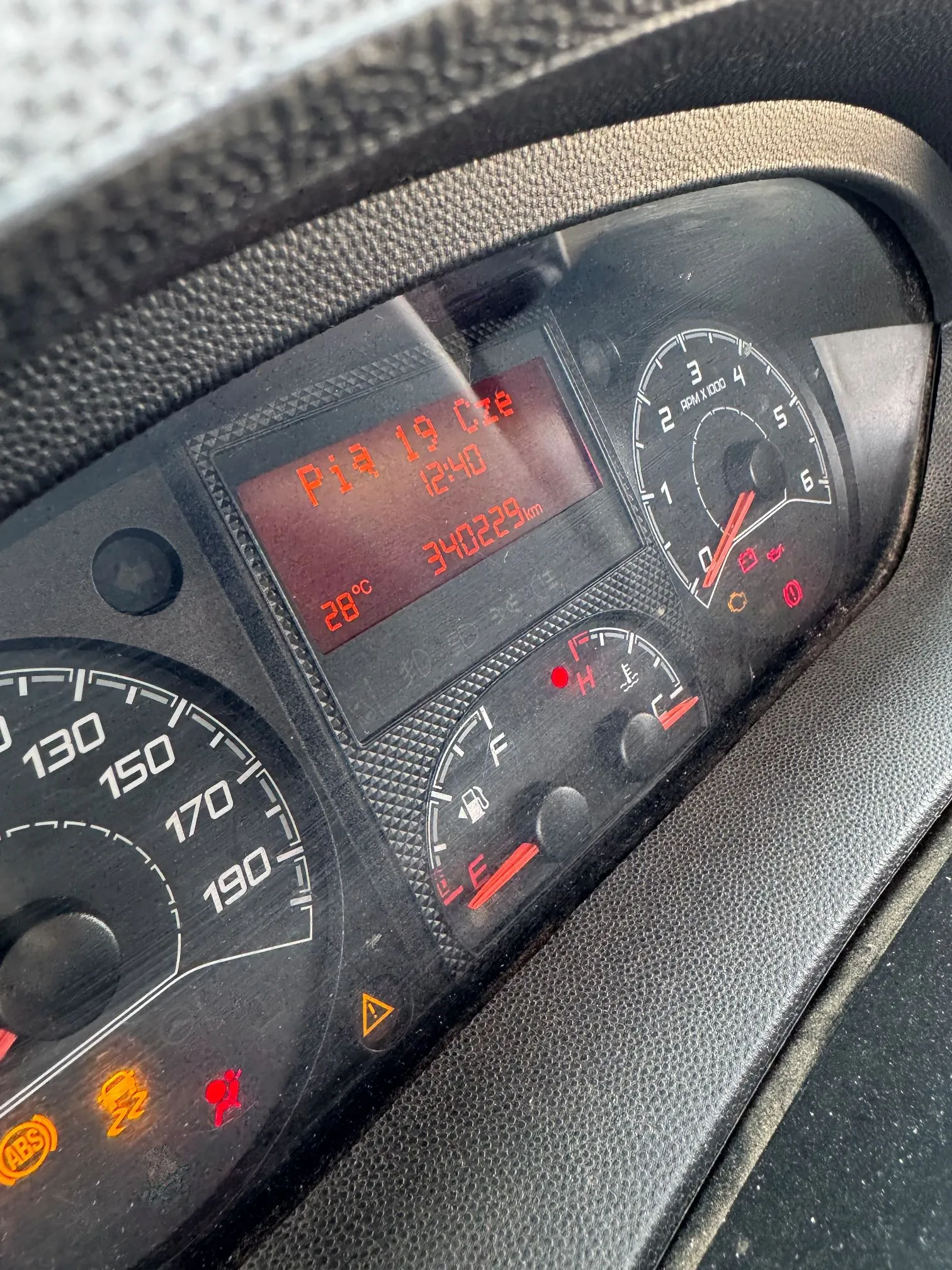 Zbliżenie na deskę rozdzielczą samochodu. Widoczny jest cyfrowy wyświetlacz z czerwonymi napisami: 'P 19 Oct', '12:40', '34022.9 km' oraz temperaturą '28°C'. Obok znajduje się analogowy wskaźnik obrotomierza z oznaczeniem 'RPM x 1000' oraz wskaźnik poziomu paliwa. W lewym dolnym rogu widoczny jest fragment prędkościomierza z wartościami 130, 150, 170, 190 oraz zapalone kontrolki ABS i inna czerwona ikona ostrzegawcza.