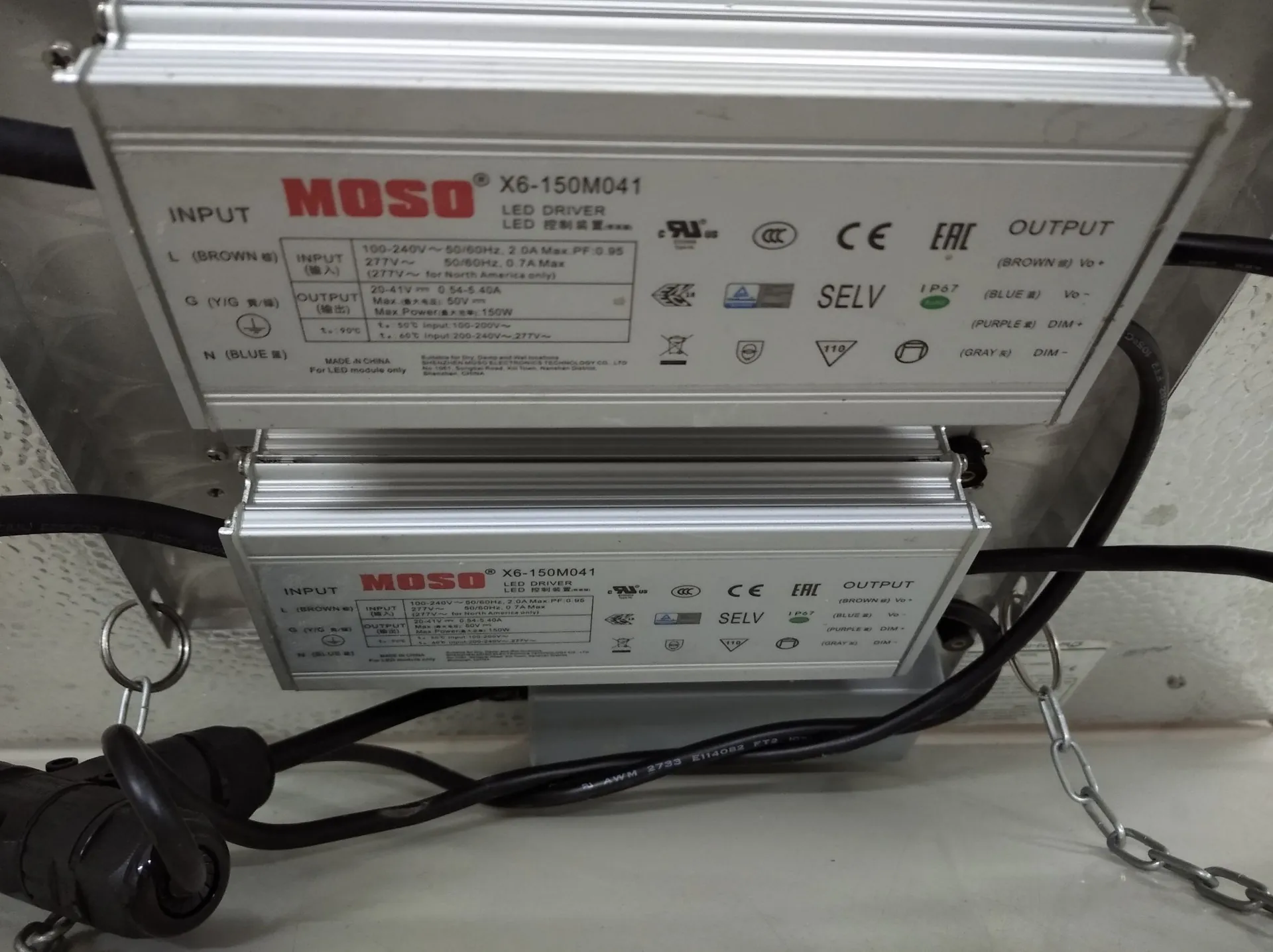 Dwa srebrne, aluminiowe zasilacze LED marki MOSO, model X6-150M041. Urządzenia mają białe etykiety z danymi technicznymi, w tym oznaczeniami wejścia (INPUT) i wyjścia (OUTPUT), certyfikatami CE, EAC oraz oznaczeniem SELV. Do zasilaczy podłączone są czarne przewody elektryczne. Urządzenia są zamontowane na jasnej powierzchni, a w dolnej części widoczny jest metalowy łańcuch oraz czarny wtyk zasilający.