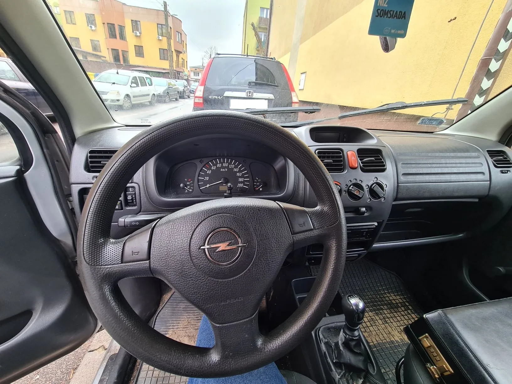 Wnętrze samochodu widziane z perspektywy kierowcy. Widoczna czarna kierownica z logo przypominającym piorun, tablica rozdzielcza z prędkościomierzem (skala do 200 km/h), deska rozdzielcza z przyciskami i wentylacjami. Po lewej stronie otwarte drzwi samochodu. Za przednią szybą widoczny czerwony samochód Honda CR-V oraz budynki mieszkalne w tle. Na prawo od szyby znak z napisem NIZ SOMSIADA. Na podłodze widoczna siateczka, a na siedzeniu pasażera czarna torebka z metalowym zamkiem.