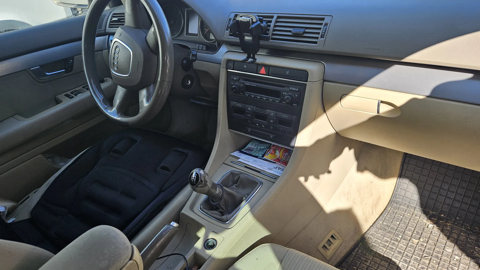 Wnętrze samochodu marki Audi z kierownicą z logo Audi, deską rozdzielczą w kolorze beżowym i szarym, centralną konsołą z radiem, klimatyzacją z napisem AUTO oraz dźwignią biegów. Na konsolie znajdują się dokumenty i gazetka. Siedzenia z czarnymi pokrowcami na beżowych poduszkach, panel drzwiowy z uchwytem i przyciskami okien, a także gumowa wykładzina na podłodze. W tle widoczne są wskaźniki prędkości i obrotomierz.