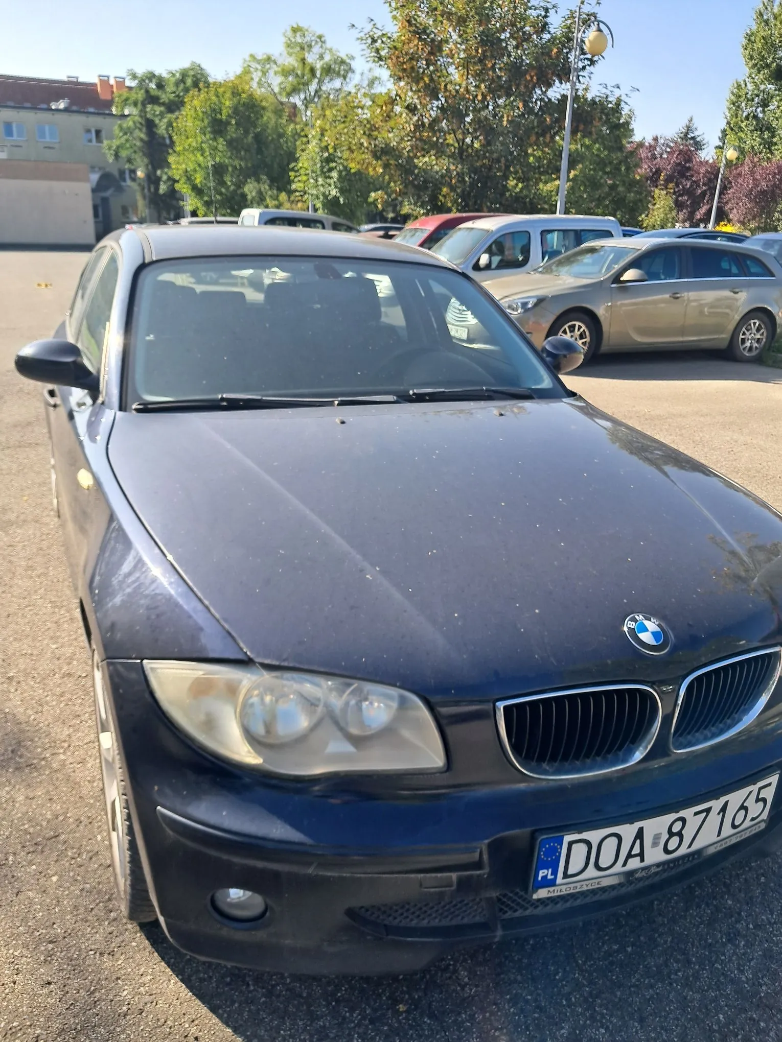 Granatowy samochód marki BMW zaparkowany na placu. Widoczny jest przód pojazdu z tablicą rejestracyjną o numerze DOA 87165. W tle znajdują się inne zaparkowane samochody, drzewa oraz fragment budynku.
