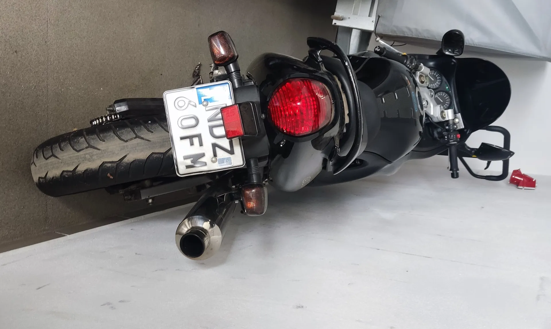 Czarny motocykl widziany od tyłu, zaparkowany przy białej ścianie. Widoczny czerwony reflektor tylny, tablica rejestracyjna z napisem LNDZ 60FM, chromowany wydech oraz liczniki na kierownicy. Podłoga z betonu, po lewej stronie fragment zakrytego pojazdu.