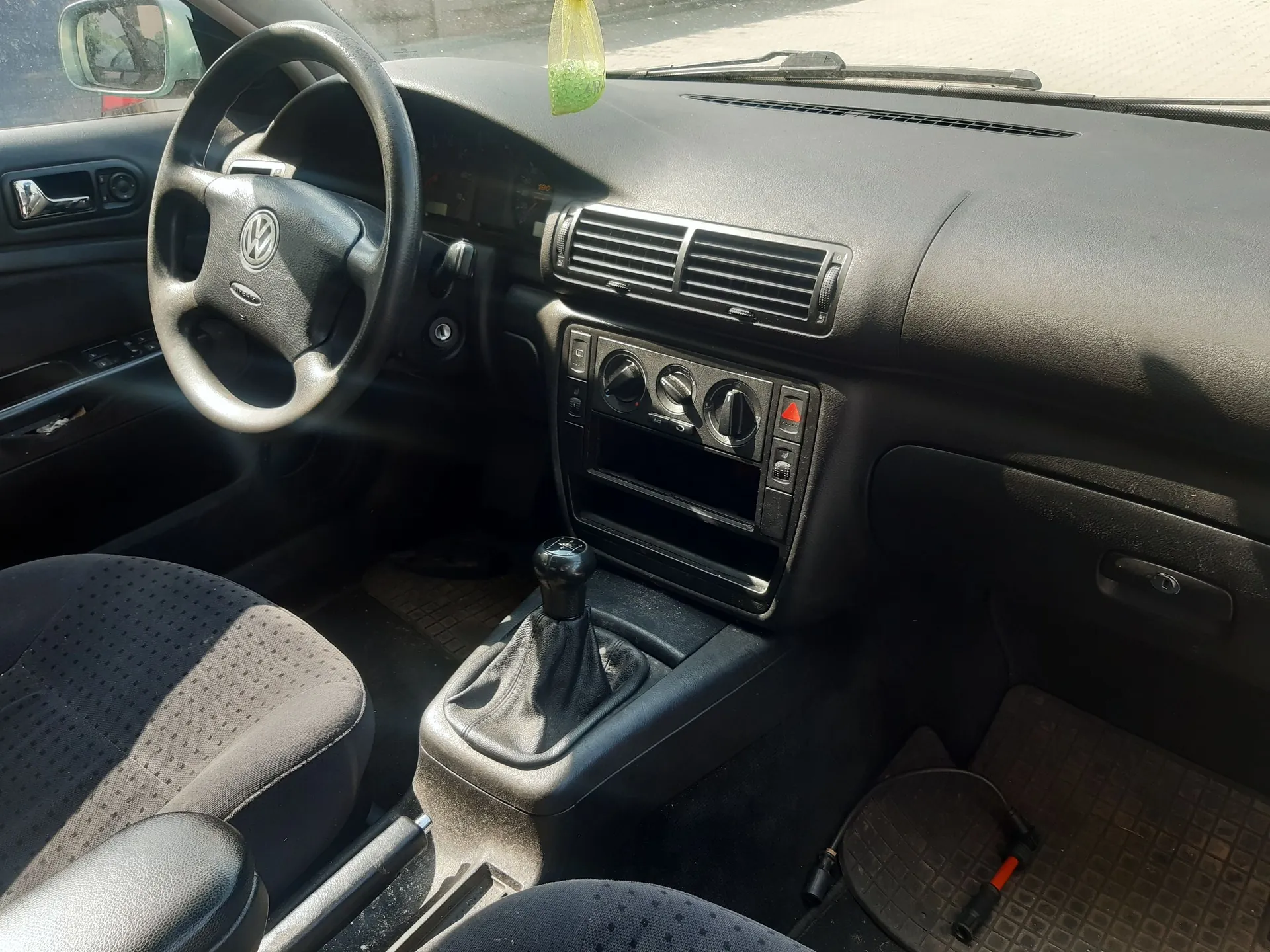 Wnętrze samochodu marki Volkswagen z perspektywy kierowcy. Widoczna jest czarna kierownica z logo VW, deska rozdzielcza z kratkami nawiewów, pokrętłami klimatyzacji oraz lewarem zmiany biegów w skórzanym mieszku. W tle widać fragmenty przednich foteli oraz lusterko wsteczne.