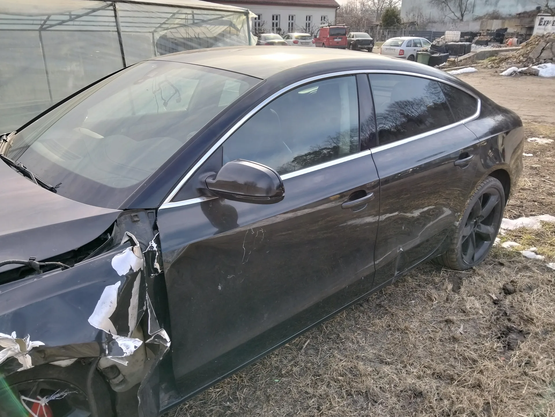 Czarny samochód osobowy z uszkodzonym przodem, w tym zdeformowaną maską i zderzakiem, zaparkowany na suchym trawniku z resztkami śniegu. W tle widoczne są inne pojazdy (czerwony van, białe i ciemne samochody), szklarnia oraz budynek z oknami. Na ziemi leżą kamienie i śmieci, a w oddali znajduje się tablica z częściowo widocznym napisem 'EXPL'.