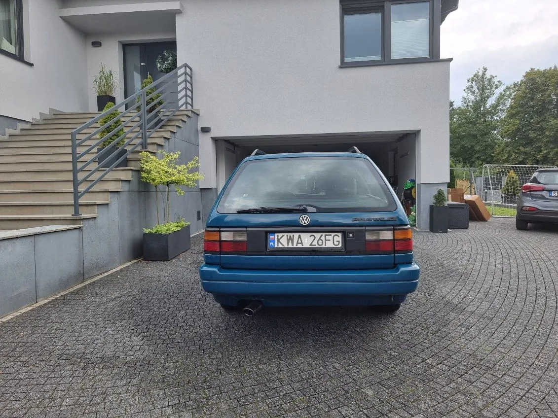 Niebieski samochód marki Volkswagen z tablicą rejestracyjną KWA 26FG zaparkowany na podjeździe przed nowoczesnym domem o szarej elewacji. Po lewej stronie schody z metalowym poręczem, donice z roślinami oraz drzwi wejściowe z wiankiem. W tle garaż, drugi samochód, bramka piłkarska i drzewa. Podjazd wyłożony kostką brukową, niebo częściowo zachmurzone.