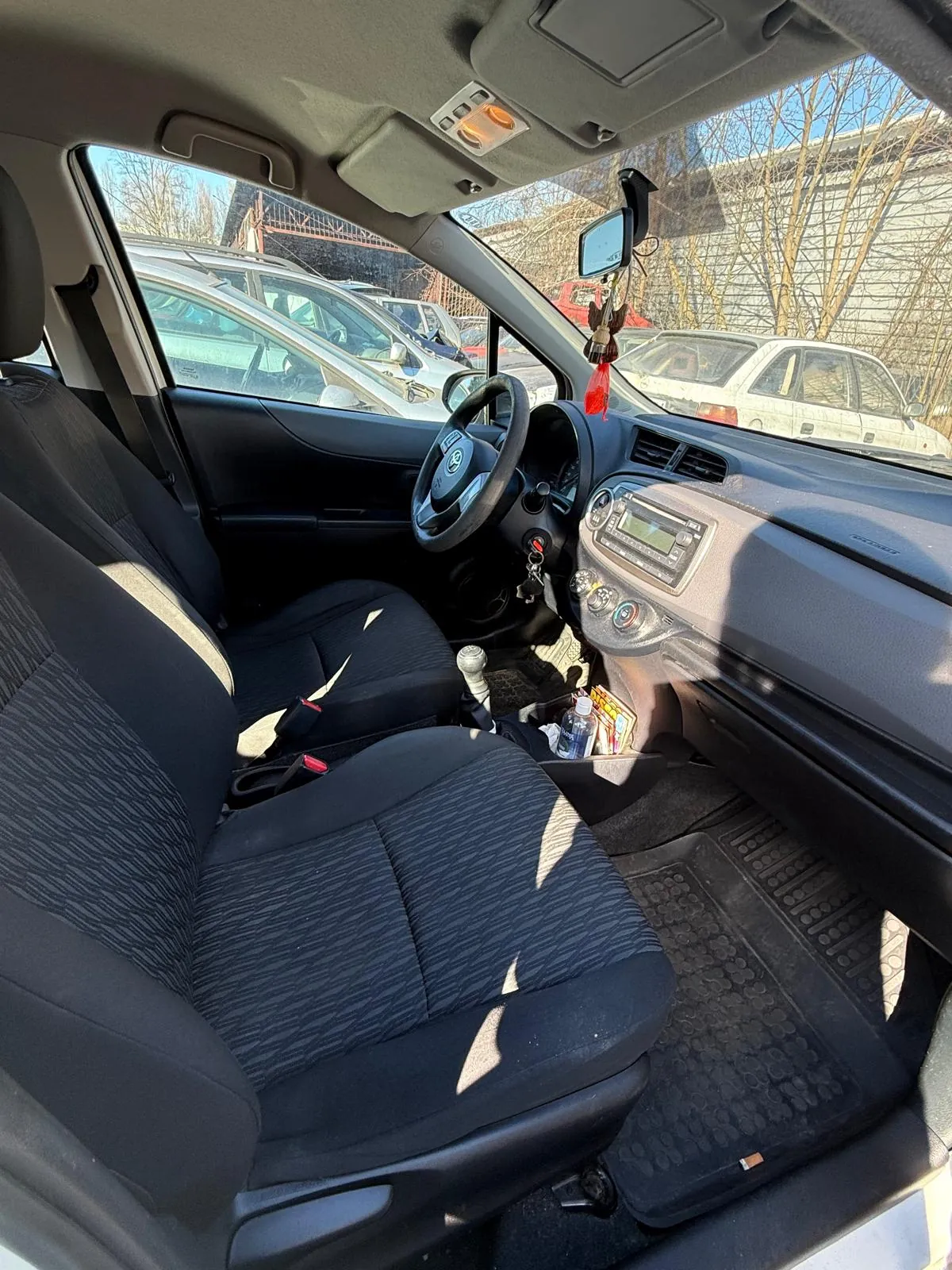 Wnętrze samochodu widziane z tyłu na przód. Czarne siedzenia z wzorzystą tapicerką, kierownica po lewej stronie, deska rozdzielcza z radioodtwarzaczem i przyciskami. W lusterku wstecznym wiszą ozdoby, w tym czerwony element. Na podłodze czarne dywaniki. Przez przednią szybę widoczne zaparkowane samochody, drzewa bez liści i budynek w tle. Jasne niebo.