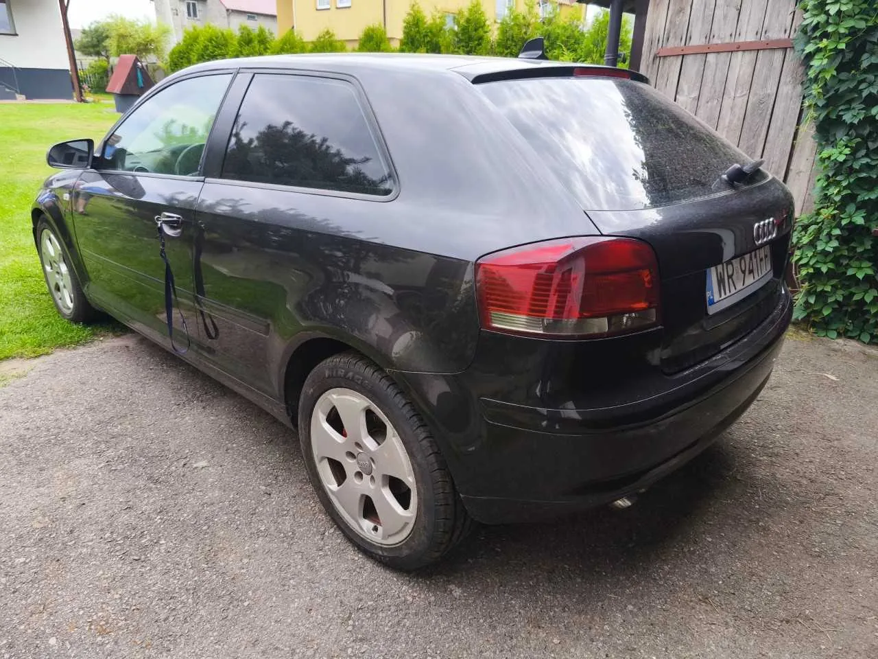 Czarny samochód marki Audi w nadwoziu hatchback, zaparkowany na żwirowym podjeździe. Widoczny jest tył i lewy bok pojazdu. Samochód ma srebrne felgi aluminiowe i polską tablicę rejestracyjną o numerze WR 941HF. W tle widać zielone krzewy, fragment drewnianego ogrodzenia oraz jasny budynek mieszkalny.