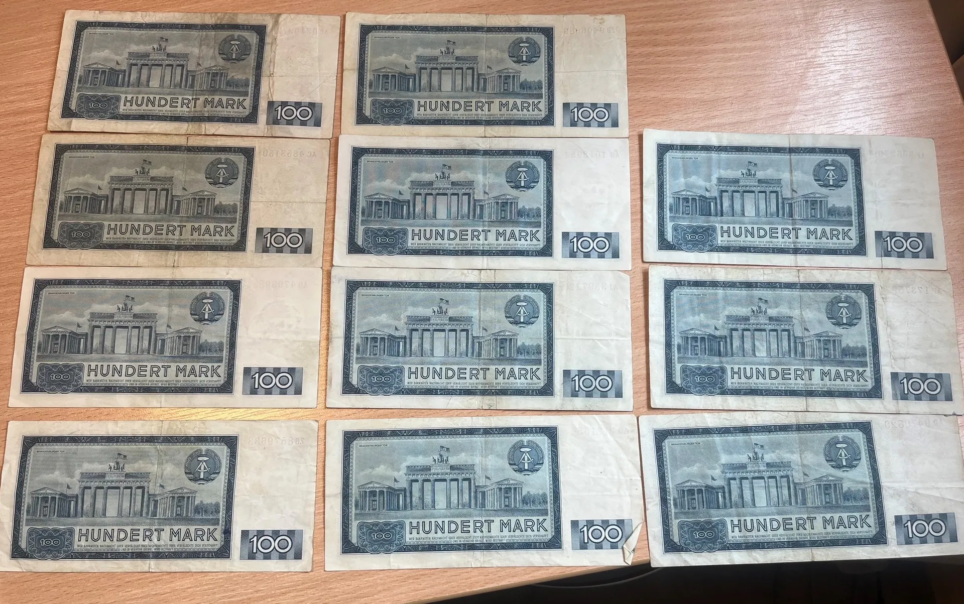 Kolekcja dwunastu niebieskawych banknotów o nominale 100 marek, ułożonych w czterech rzędach po trzy sztuki na drewnianym blacie. Na każdym banknocie widnieje napis „HUNDERT MARK”, liczba „100” oraz grafika przedstawiająca Bramę Brandenburską w Berlinie.