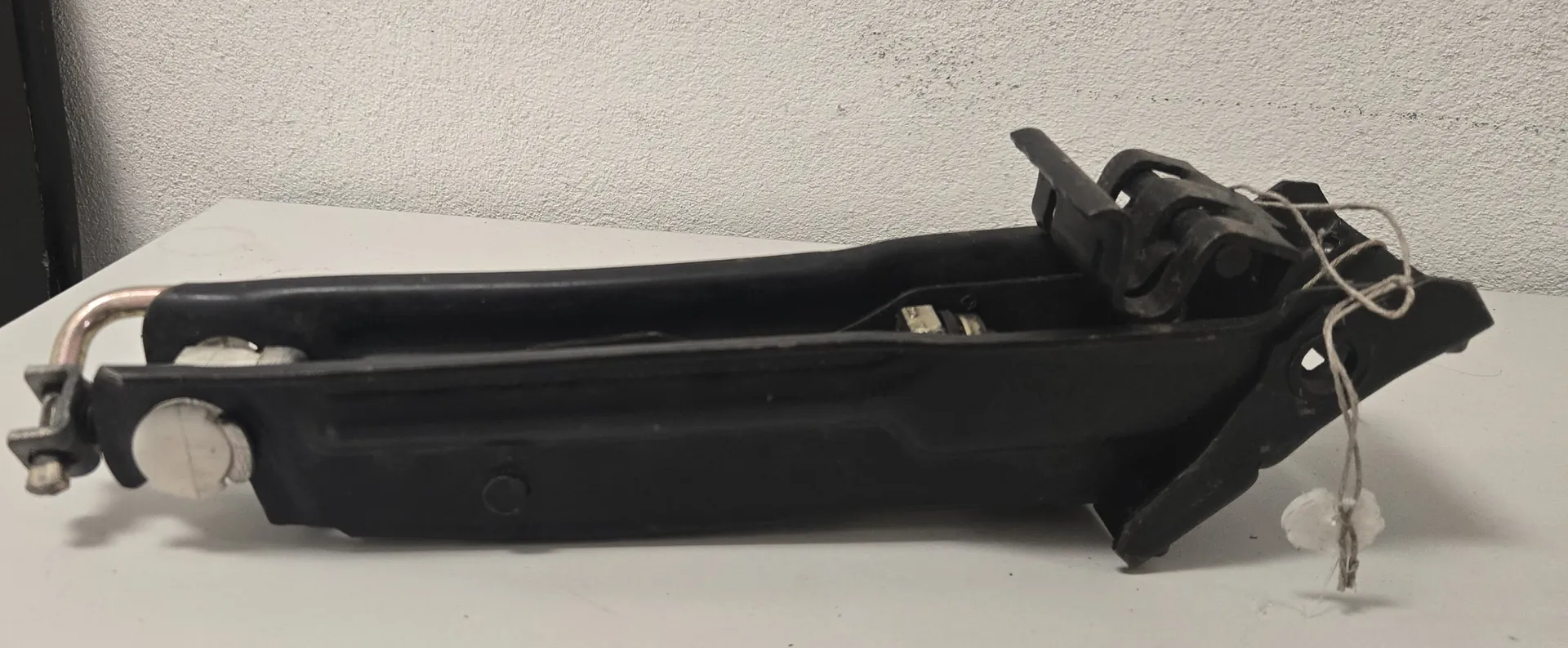 Czarna, plastikowa część mechaniczna, prawdopodobnie element klamki lub mechanizmu blokady z samochodu, leżąca poziomo na białym blacie. Przedmiot posiada metalowe elementy mocujące po lewej stronie oraz sznurek zawiązany wokół prawego końca.