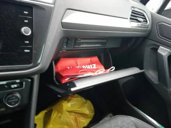 Wnętrze samochodu z widoczną deską rozdzielczą, systemem rozrywki z przyciskami 'RADIO', 'CAR', 'MEDIA' po lewej stronie. Otwarty schowek przedni zawiera czerwoną torbę z napisem 'Ziho' oraz żółtą torbę plastikową poniżej. Wnętrze wykończone szarym materiałem.