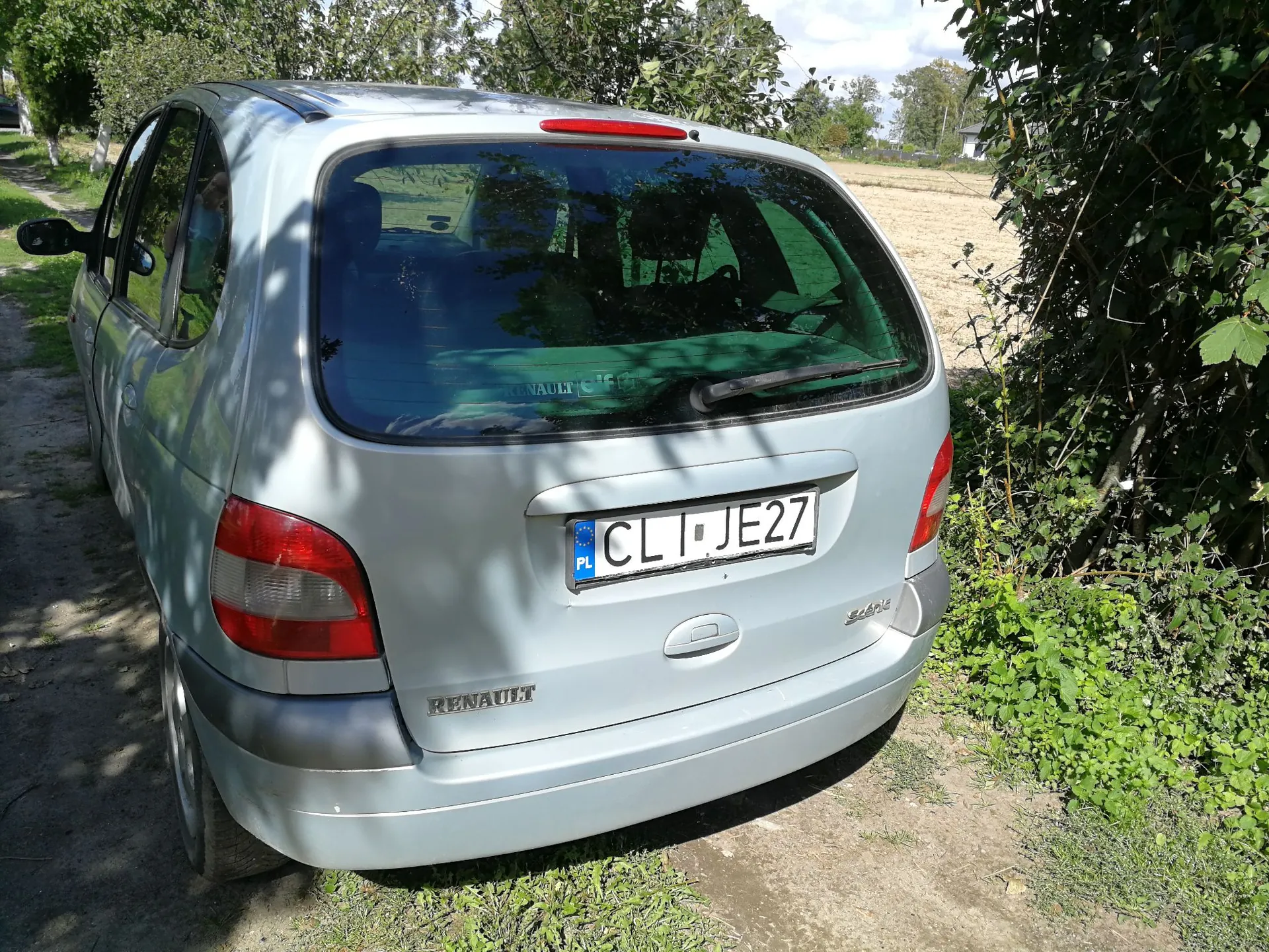 Srebrny samochód marki Renault Scenic zaparkowany na piaszczystej drodze obok zielonych krzewów i pola. Widoczny jest tył pojazdu z polską tablicą rejestracyjną o numerze CL JE27 oraz napisem RENAULT na klapie bagażnika.