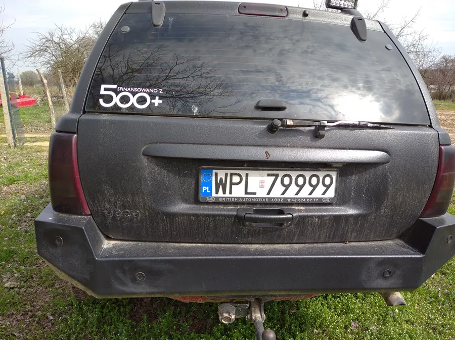 Tył czarnego samochodu marki Jeep z zabrudzoną karoserią. Na szybie tylnej naklejka z napisem "500+ SPINANSOWANO Z". Tablica rejestracyjna z białym tłem i czarnym tekstem "WPL 79999", po lewej stronie niebieski pas z napisem "PL" oraz napisem "BRITISH AUTOMOTIVE ŁÓDŹ" pod tablicą. Samochód stoi na trawie, w tle drzewa bez liści i ogrodzenie. Niebo częściowo zachmurzone.