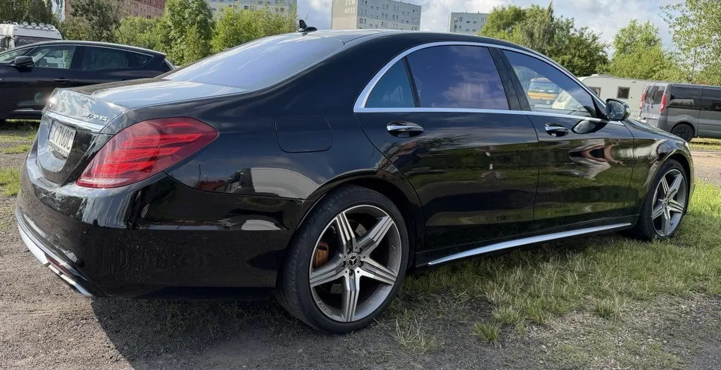 Czarny samochód osobowy marki Mercedes-Benz zaparkowany na trawiasto-żwirowym podłożu. Widok z tyłu i z boku. Auto posiada srebrne felgi z pięcioramiennym wzorem. W tle widoczne są inne zaparkowane pojazdy, drzewa oraz bloki mieszkalne.