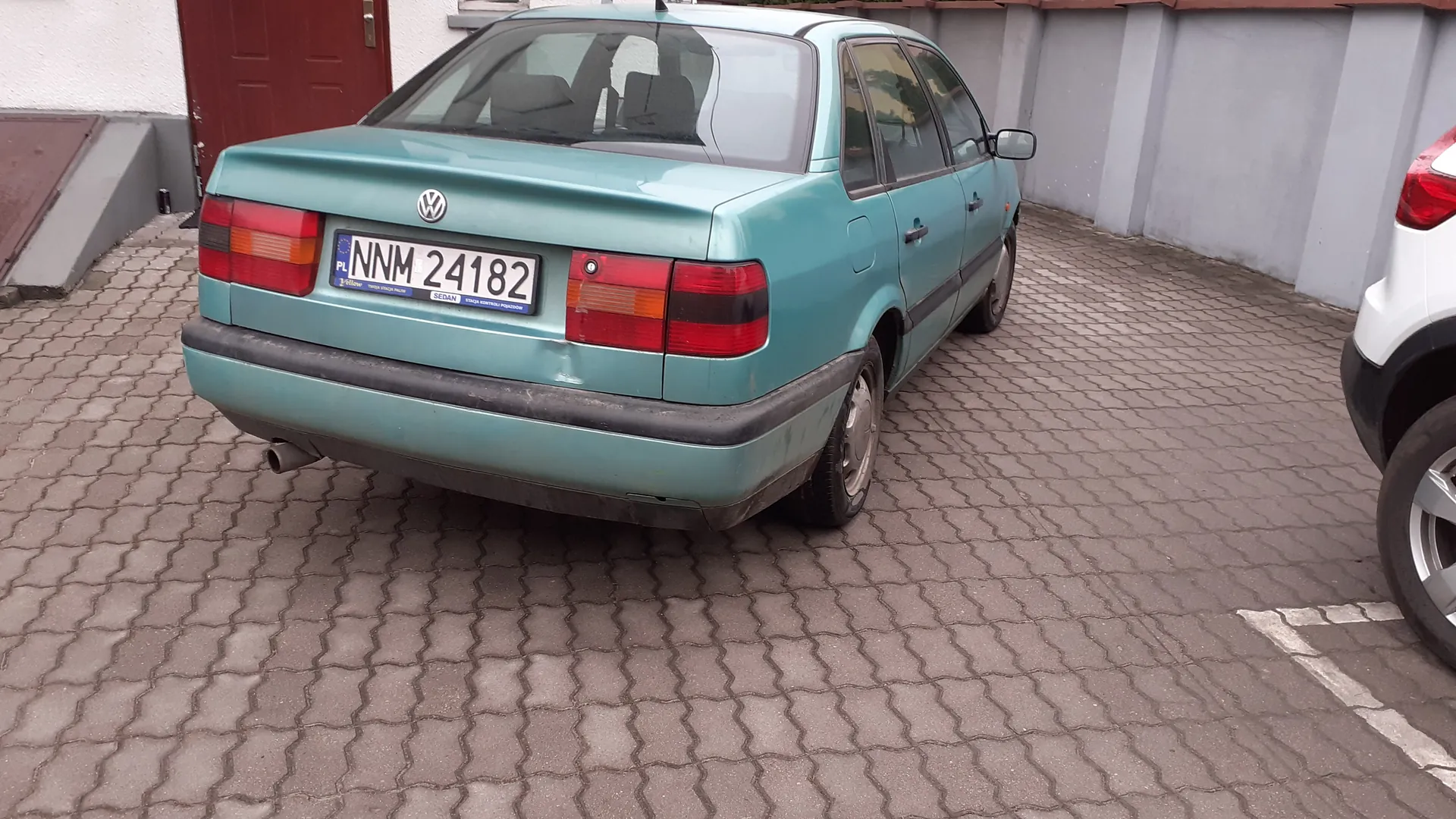 Zdjęcie przedstawia tył jasnoniebieskiego samochodu marki Volkswagen, zaparkowanego na brukowanej powierzchni. Widoczna jest tablica rejestracyjna z napisem PL NNM 24182 oraz logo Volkswagena na bagażniku. Po lewej stronie tła znajduje się czerwone drzwi i biała ściana, po prawej fragment białego samochodu. Samochód ma czerwone światła tylna i czarny zderzak. W tle widoczne są szare ściany budynku.