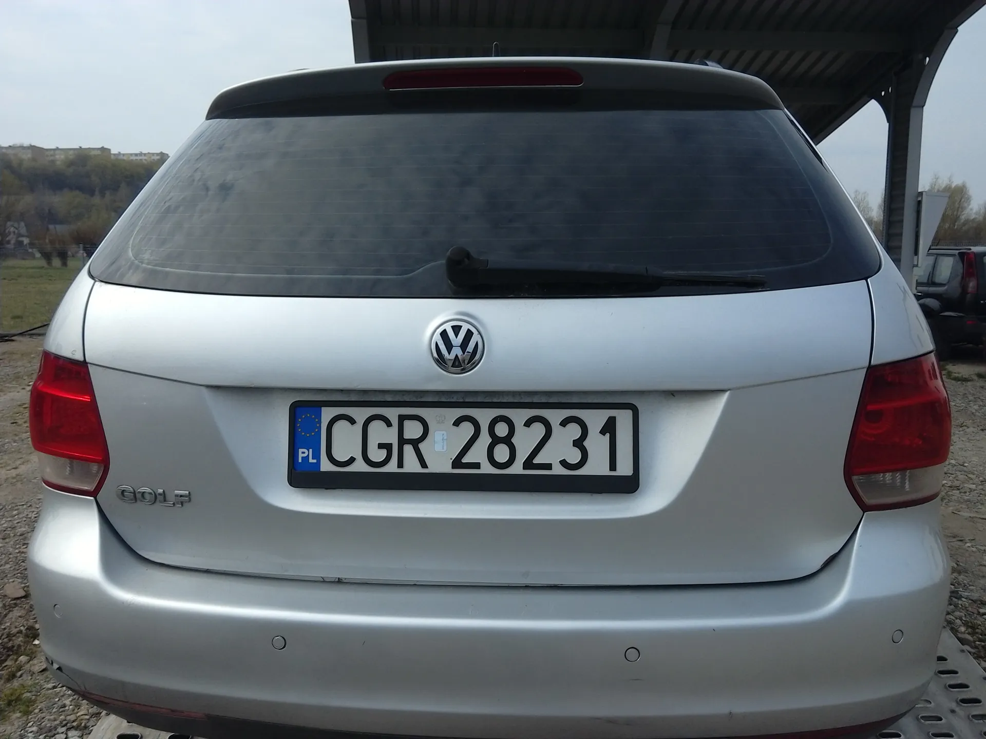 Tył srebrnego samochodu marki Volkswagen Golf. Widoczna tablica rejestracyjna z napisem 13282 RG C oraz niebieskim pasem z napisem PL i symbolem Unii Europejskiej po prawej stronie. Na bagażniku umieszczony emblemat VW oraz napis GOLF po lewej stronie. Samochód zaparkowany na żwirowym podłożu, w tle fragment budynku i niebo.