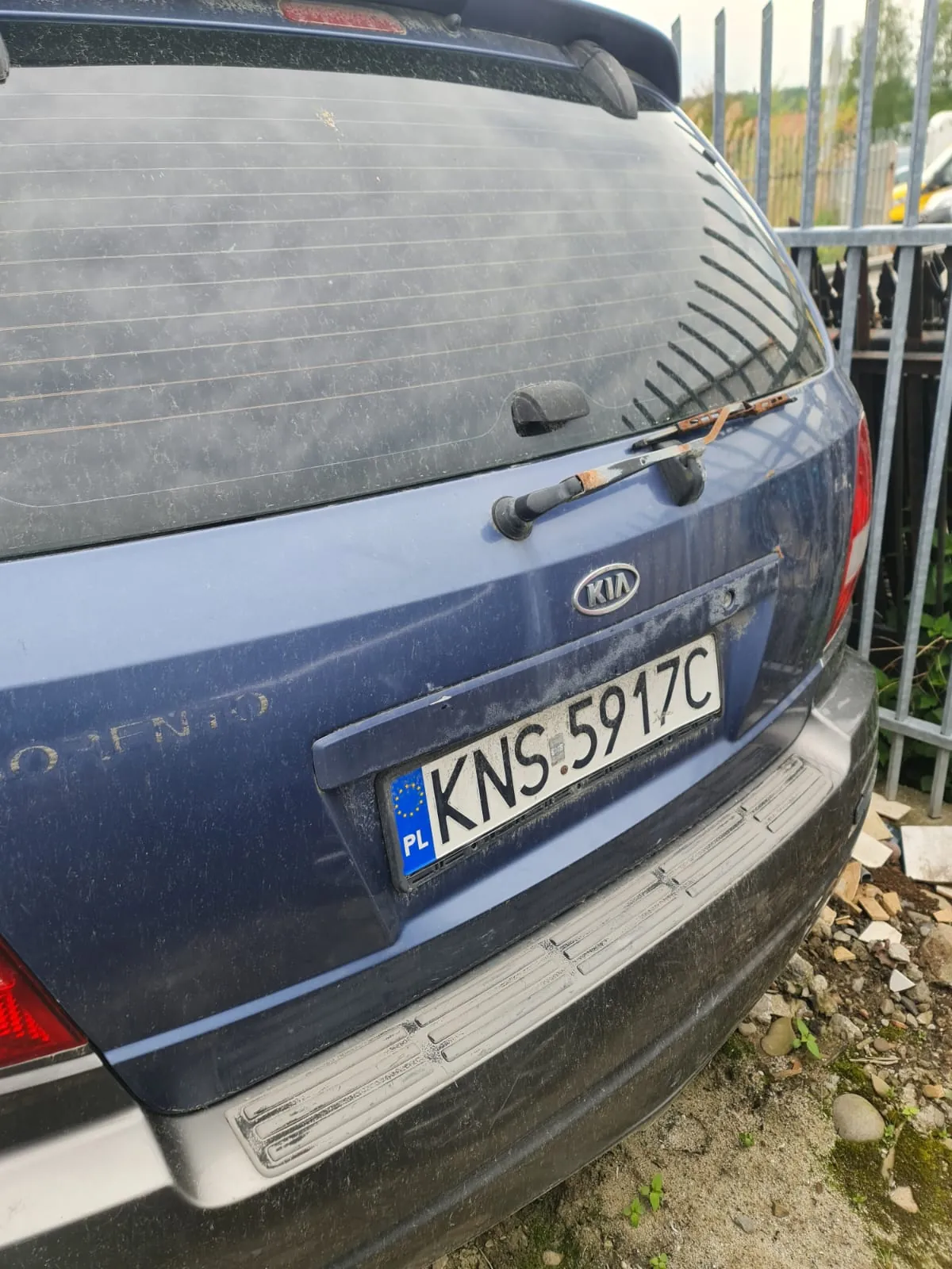 Tył niebieskiego samochodu marki Kia z widocznym logo i napisem "ORENTO" na bagażniku. Tablica rejestracyjna z białym tłem, niebieskim pasem z napisem "PL" i symbolem UE, oraz czarnym tekstem "KNS 5917C". Samochód jest zabrudzony, a na tle widoczne są metalowe ogrodzenie, kamienie i ziemia. W tle również fragmenty innych pojazdów i roślinność.