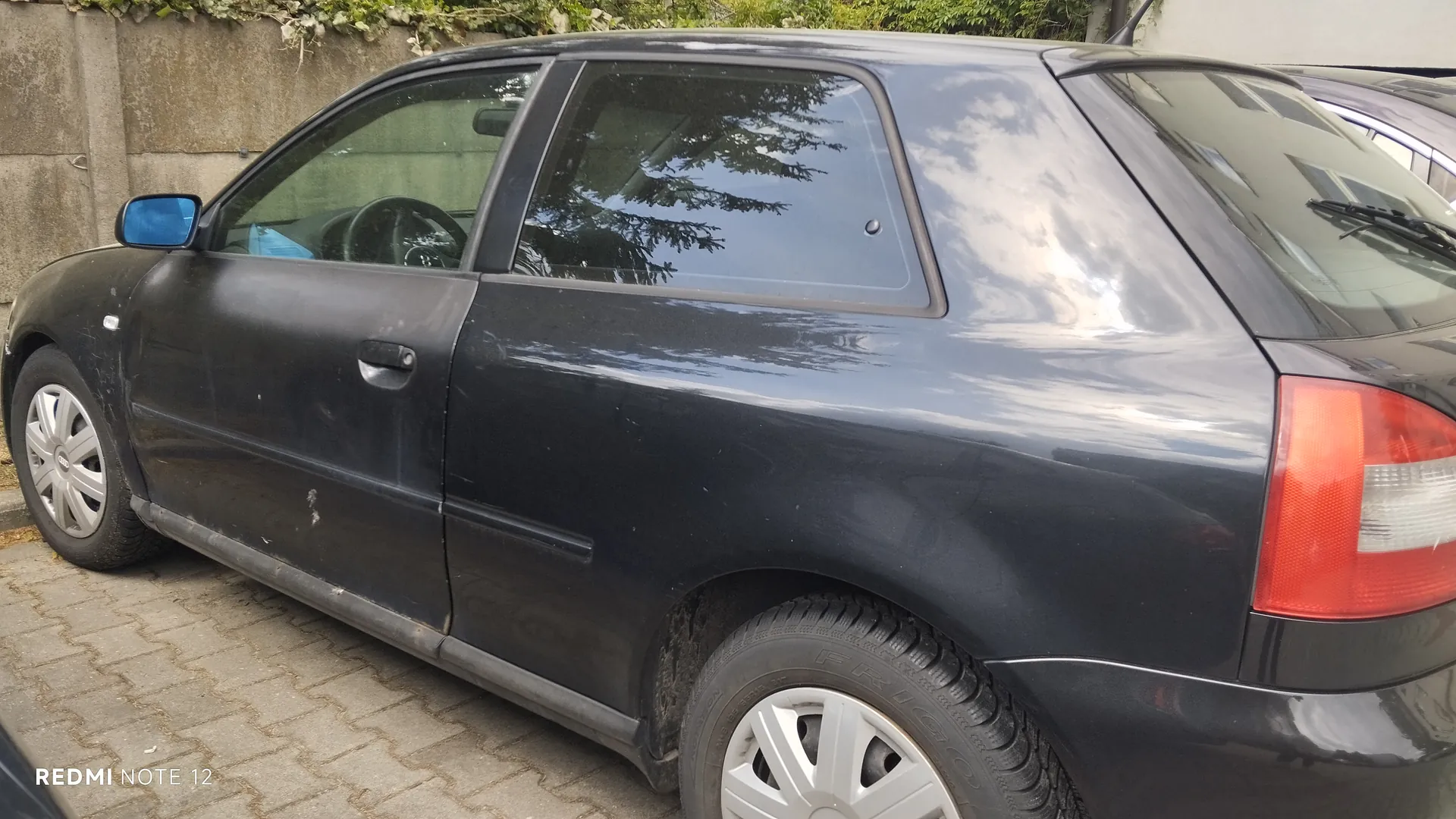 Czarny samochód osobowy typu hatchback zaparkowany na brukowanej powierzchni. Widoczne są boczne drzwi, tylne światło oraz felgi z srebrnymi dyskami. W tle betonowa ściana z zielonymi pnączami oraz fragment innego pojazdu. Na dole zdjęcia widoczny napis 'REDMI NOTE 12'. Samochód ma ślady brudu na karoserii, a w szybach odbijają się drzewa i niebo.
