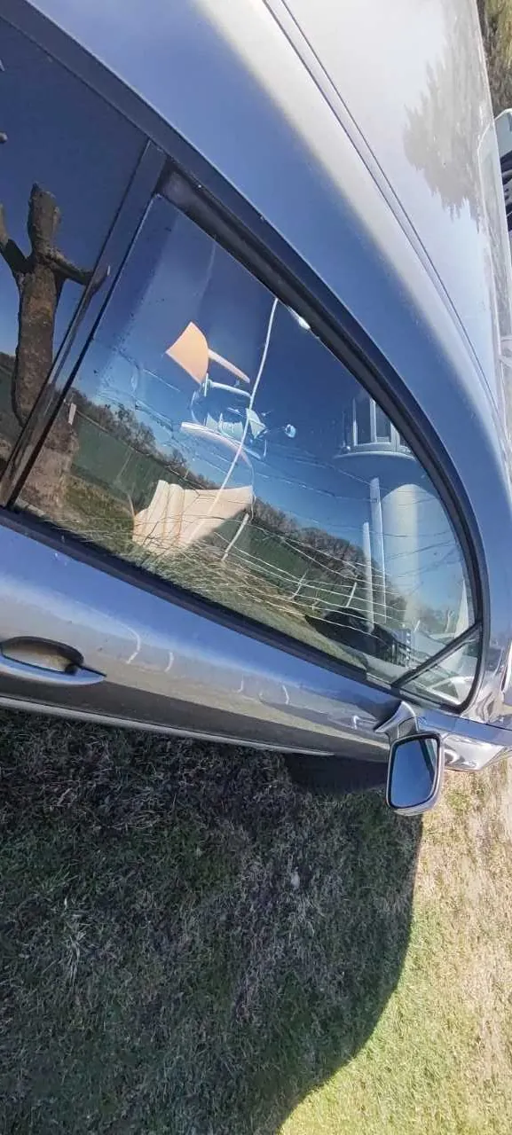 Srebrny samochód z pękniętym szybą pasażerską. Widoczne pęknięcia na szybie, boczne lusterko, uchwyt drzwiowy oraz odbicie w szybie przedstawiające elementy otoczenia. Samochód zaparkowany na trawie.