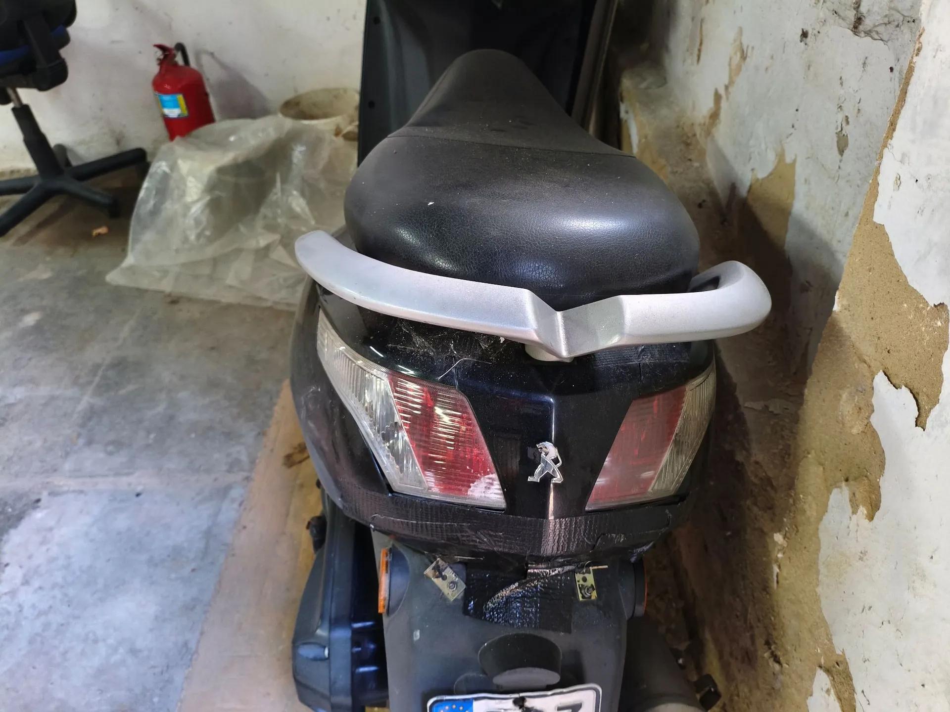 Widok z tyłu na czarny skuter marki Peugeot. Widoczne jest czarne siedzenie, srebrny uchwyt dla pasażera oraz tylna lampa z logo lwa. Poniżej lampy znajduje się fragment tablicy rejestracyjnej. Skuter stoi w pomieszczeniu z odniszczoną ścianą, a w tle widać gaśnicę i plastikową folię.