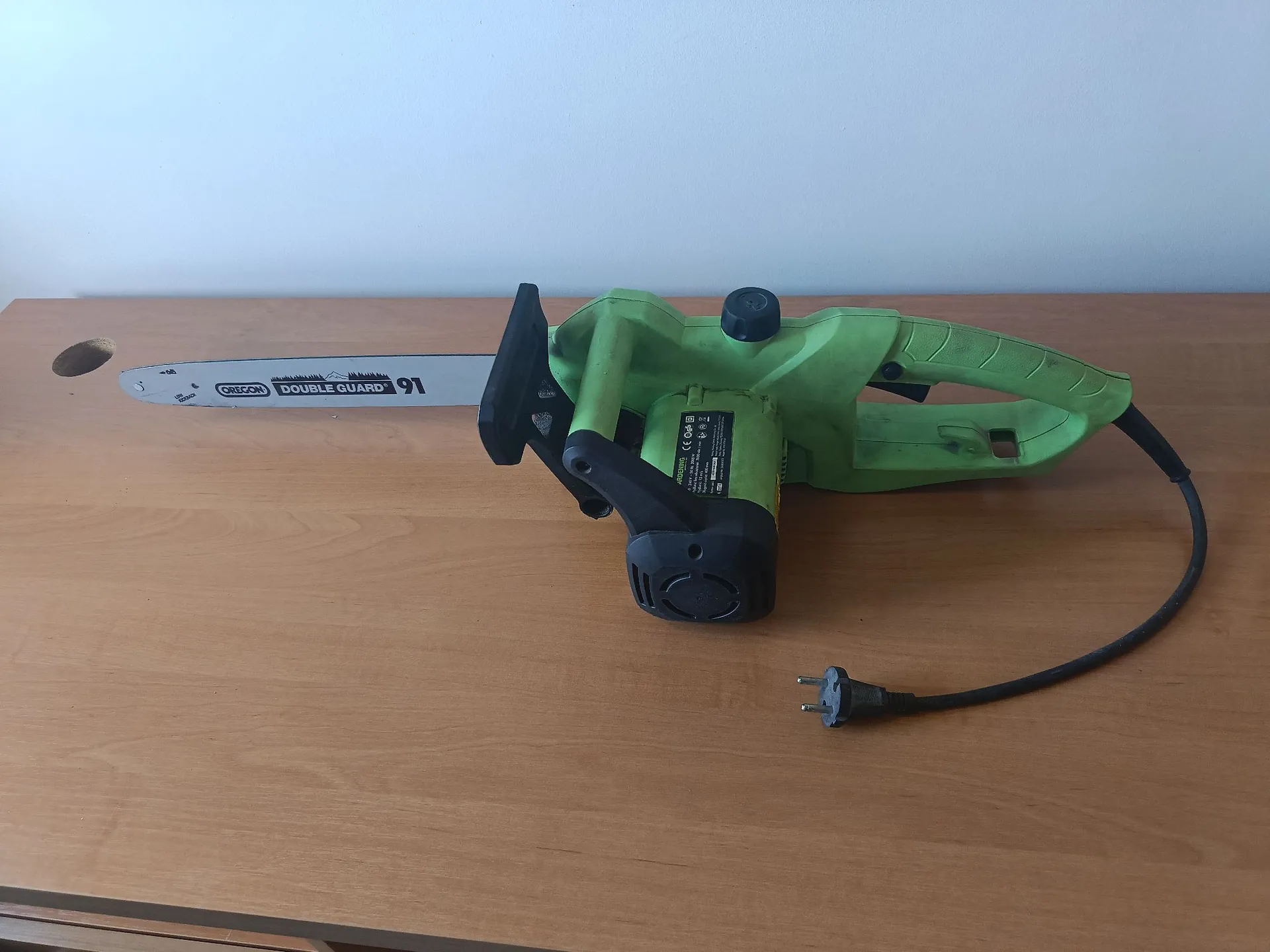 Zielona piła łańcuchowa elektryczna leżąca poziomo na drewnianym blacie. Na prowadnicy piły widnieje napis 'OREGON DOUBLE GUARD 91'. Urządzenie posiada czarny kabel z wtyczką oraz czarną obudowę silnika z tabliczką znamionową.