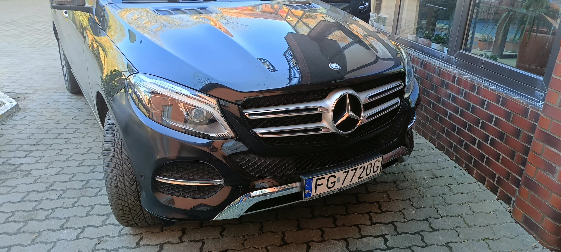 Czarny samochód Mercedes-Benz SUV zaparkowany na brukowanej powierzchni. Widoczna przód pojazdu z charakterystyczną atrapą chłodnicy z logo trójgwiazdy, reflektorem i tablicą rejestracyjną FG 7720G z niebieskim pasem PL po lewej stronie. Po prawej stronie obrazu ceglana ściana i duże okno z roślinami w doniczkach. Odbicia na błyszczącej karoserii wskazują na słoneczny dzień.