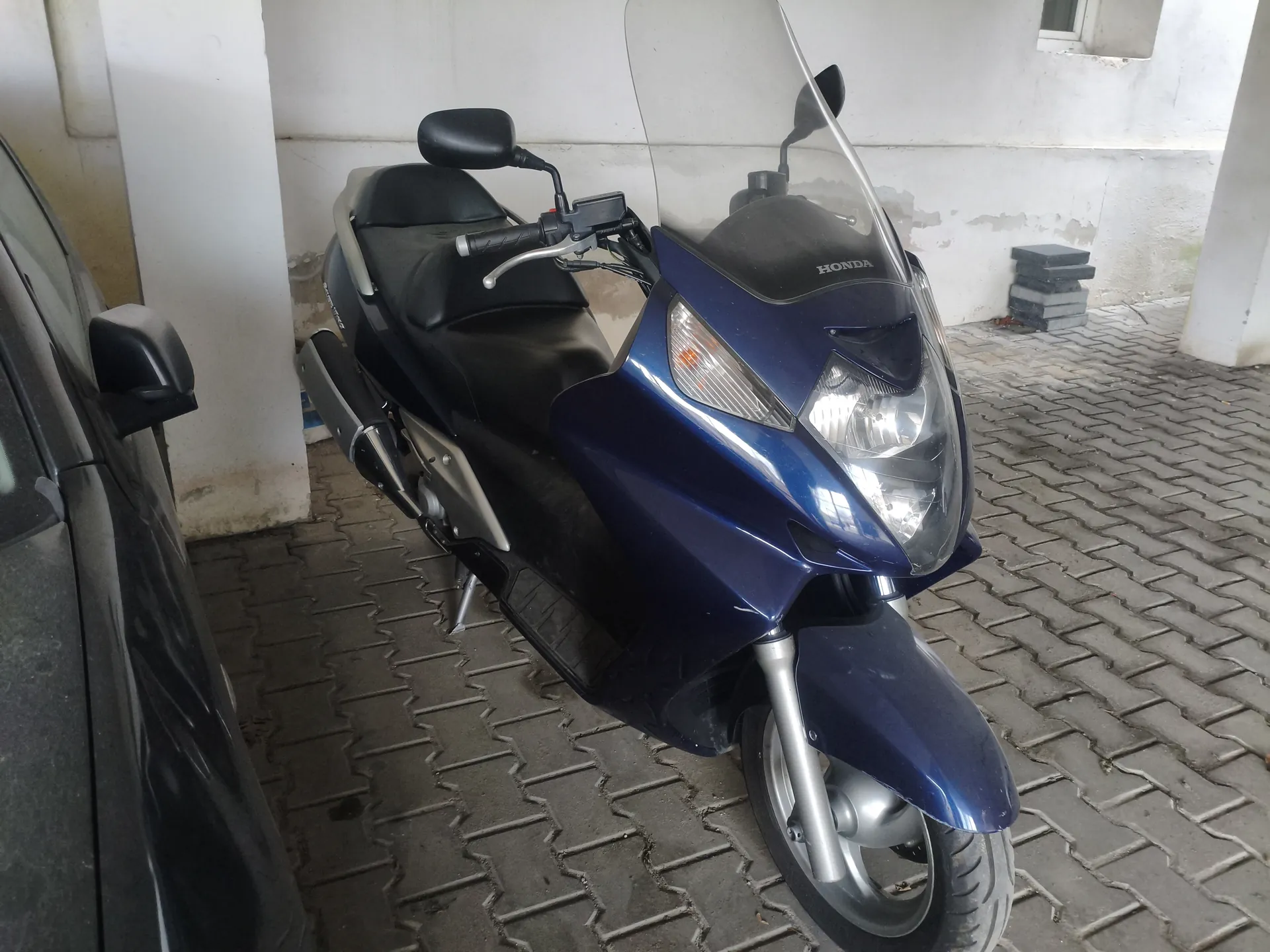 Niebieski skuter marki Honda z czarnym siedzeniem i przednią szybą, zaparkowany na brukowanej powierzchni. Po lewej stronie widoczna część ciemnego samochodu. Tło stanowi biała ściana z oznakami zużycia oraz stos czarnych przedmiotów. Na przedniej części skutera widoczny napis HONDA.