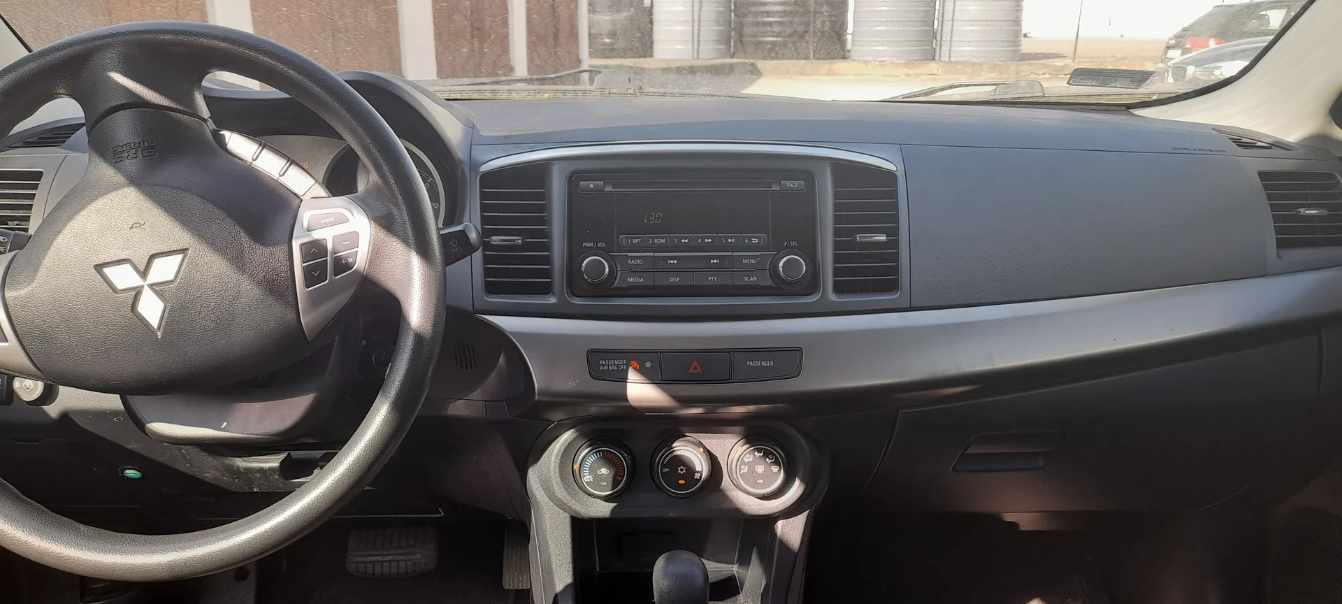 Wnętrze samochodu z widocznym kierownicą z logo Mitsubishi, centralnym panelem z odtwarzaczem muzycznym wyświetlającym godzinę 1:30, przyciskami sterującymi (RADIO, MEDIA, MENU), wentylacjami, pokrętłami klimatyzacji oraz dźwignią zmiany biegów. Przez przednią szybę widoczne są budynki i beczki na zewnątrz.