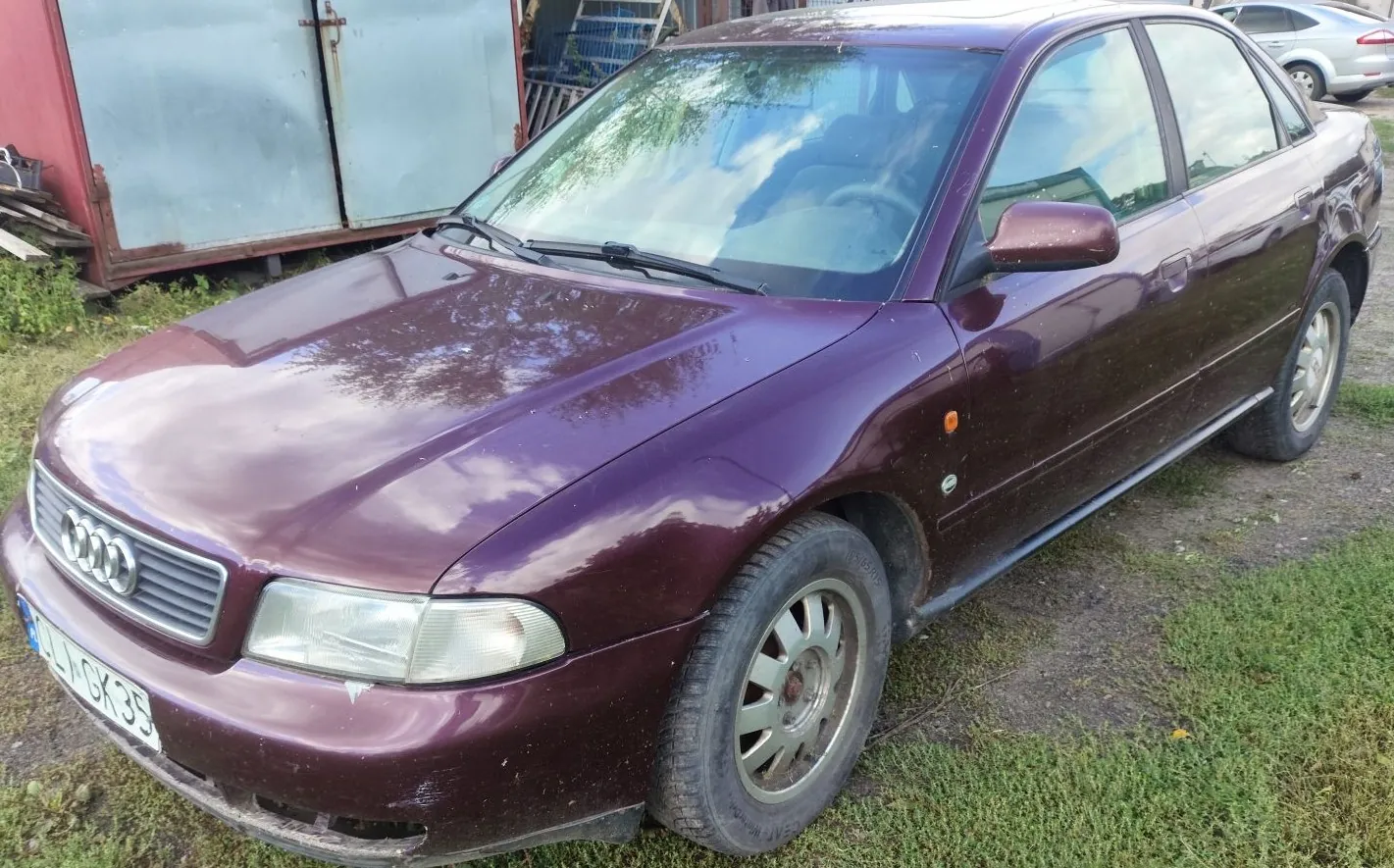 Bordowy samochód marki Audi zaparkowany na trawiasto-żwirowym podłożu. Widoczny jest przód i prawy bok pojazdu. Samochód ma tablicę rejestracyjną zaczynającą się od liter LI GK35. W tle znajduje się metalowy kontener oraz fragment innego, srebrnego samochodu.